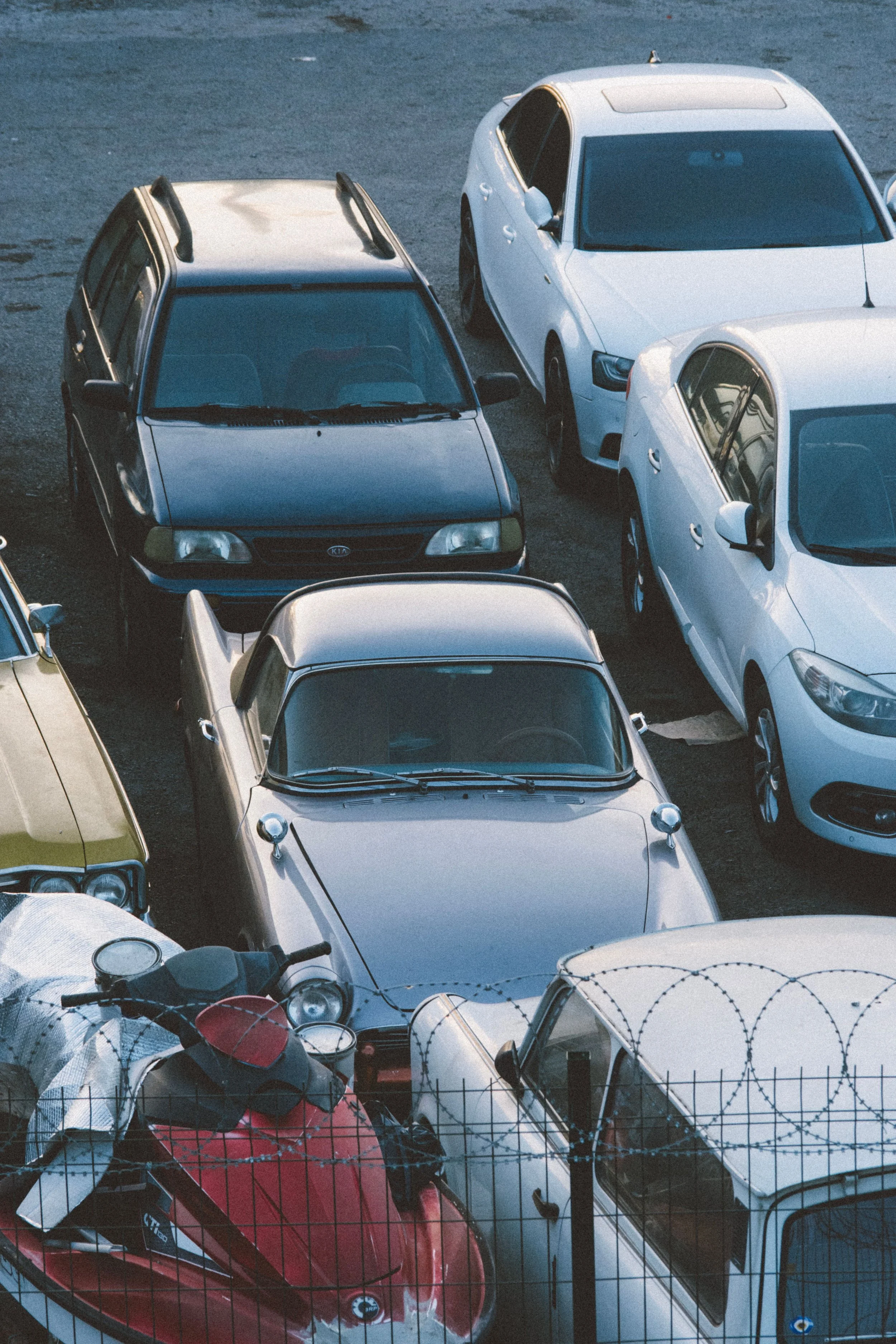 A parking lot with several cars parked, including a black Kia, white sedans, and a yellow car, with a motorcycle and a red scooter in the foreground behind a barbed wire fence.