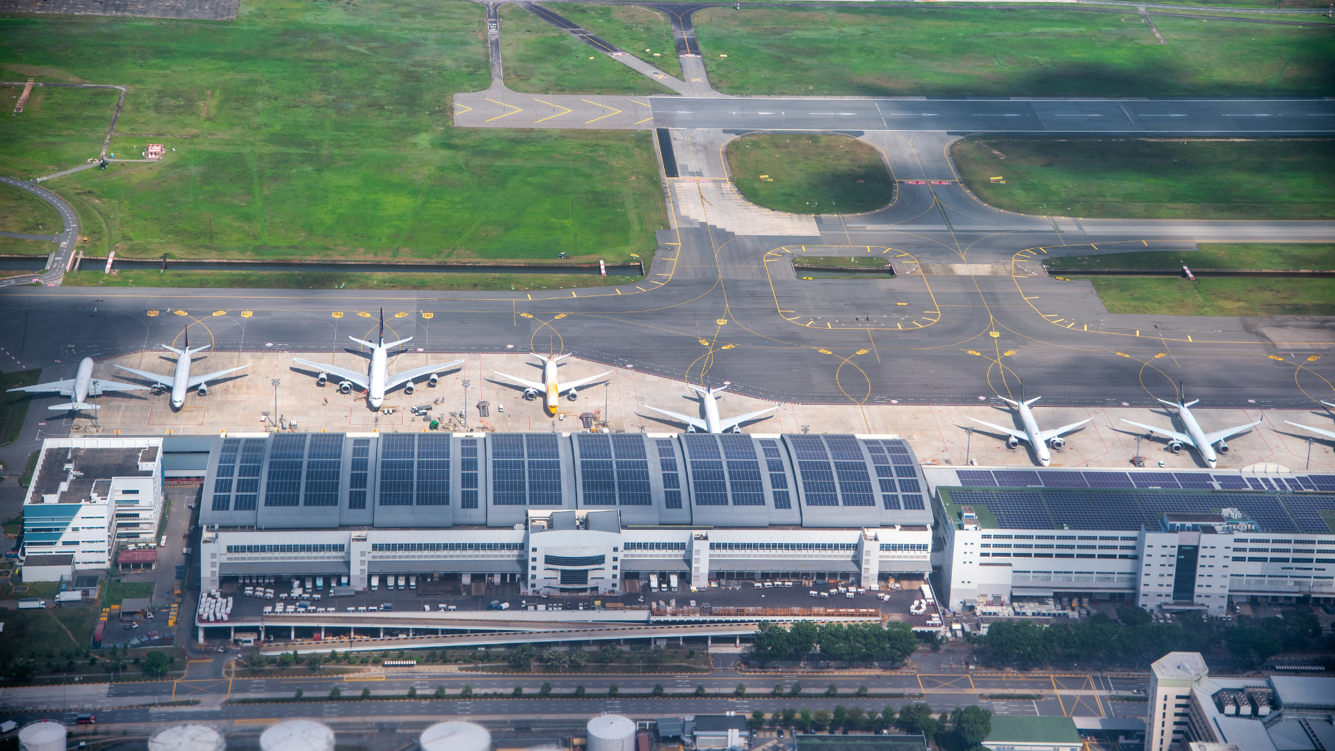 Aerial view of an airport with six parked airplanes at the gate, surrounding airport buildings, and taxiways leading to runways, with green grass areas and roads outside the airport complex.
