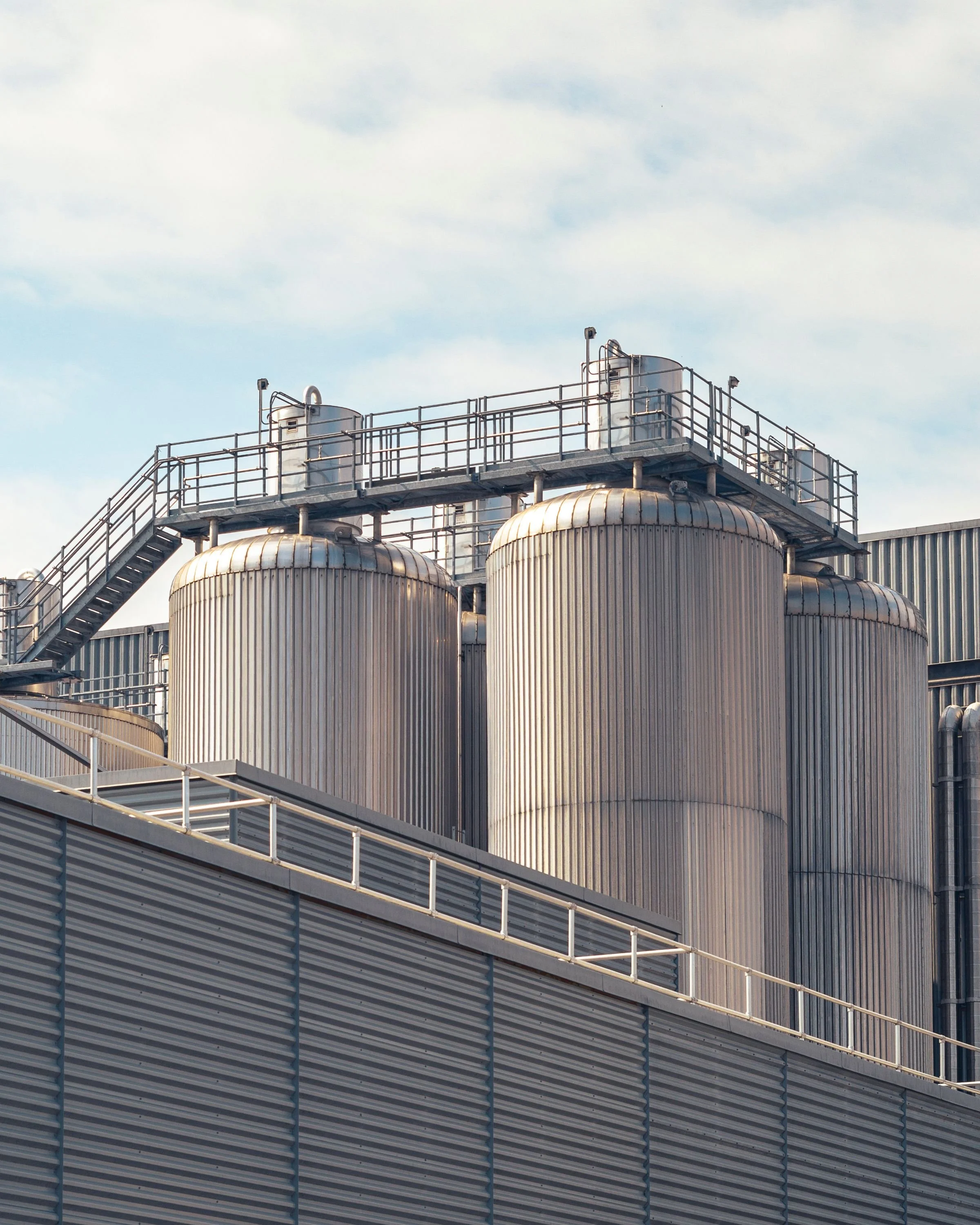 Industrial facility with large metallic tanks and overhead walkways against a partly cloudy sky.