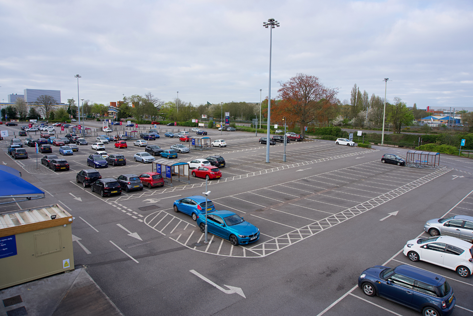 A mostly empty parking lot with a few cars parked and some empty spaces, surrounded by trees and buildings in the background, under a cloudy sky.