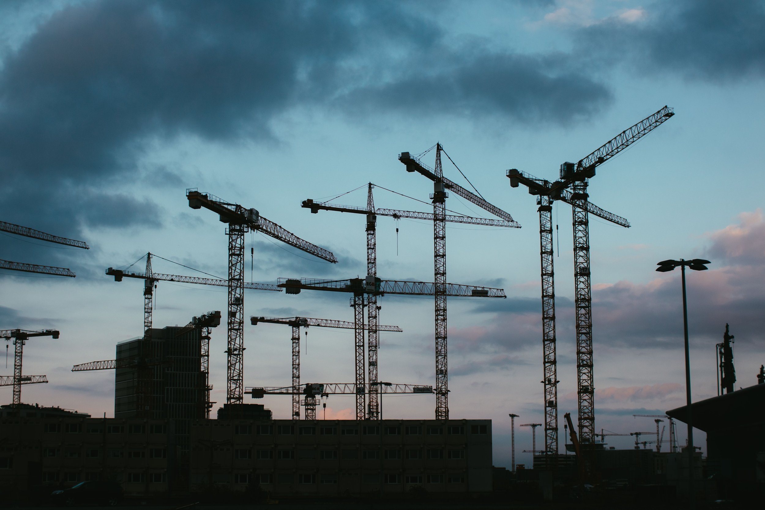 silhouettes of construction cranes against a cloudy evening sky over a cityscape.