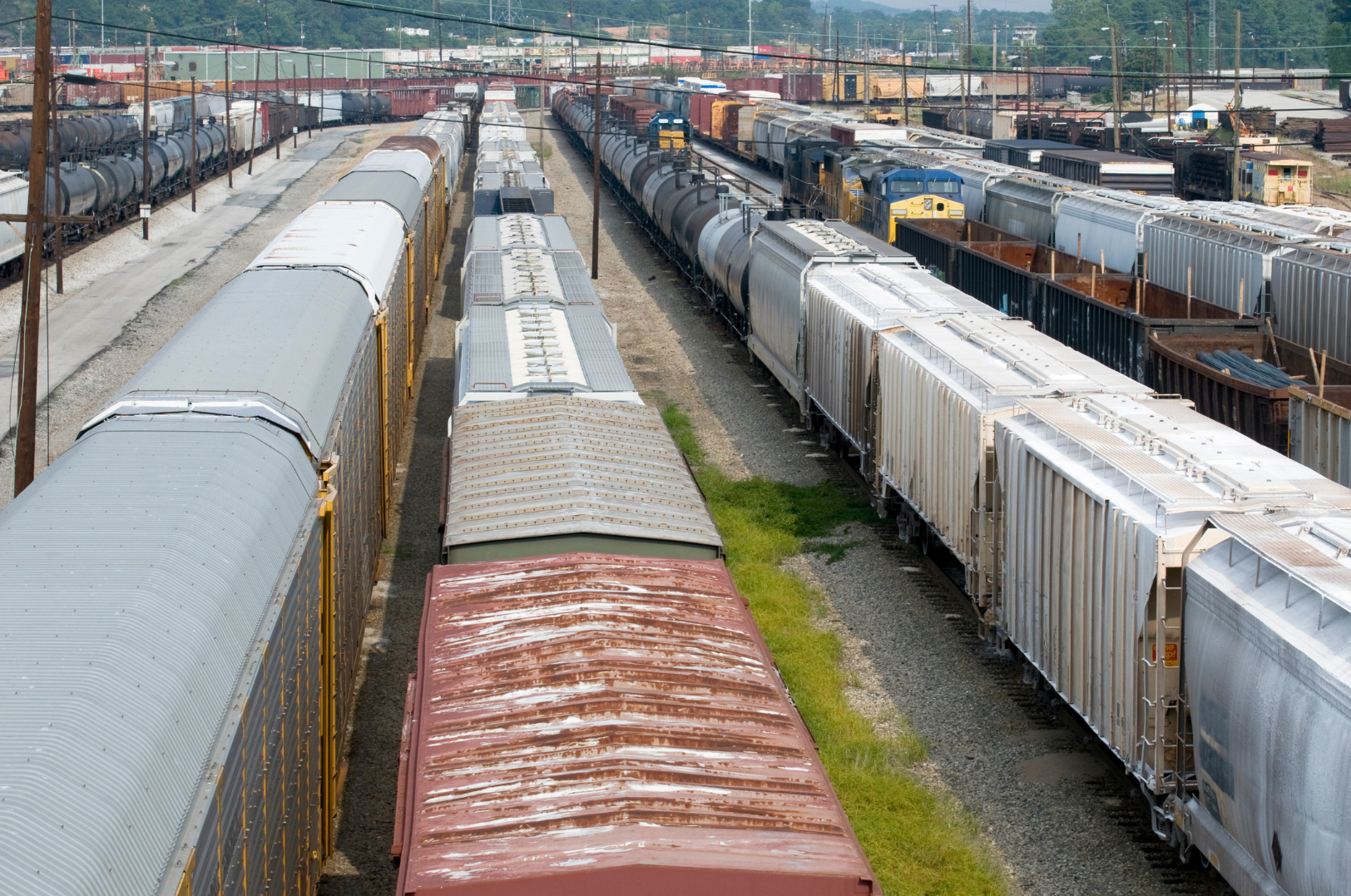 A large train yard with multiple freight trains on parallel tracks, including tank cars, boxcars, and flatcars, with some trains appearing rusty and weathered, and a train engine in the background, set against a backdrop of green mountains and industrial buildings.