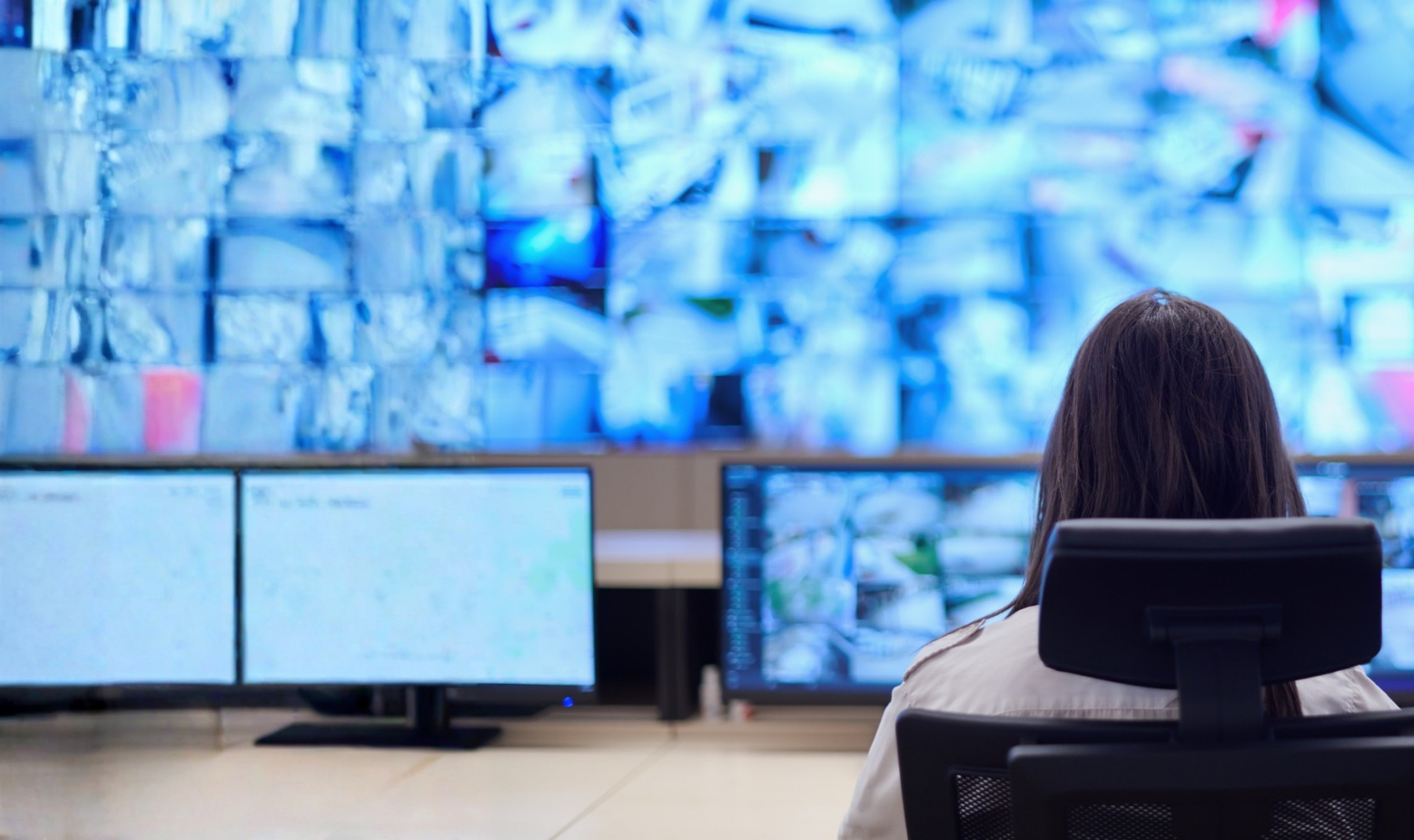 A person sitting at a desk with multiple monitors, observing a large digital display wall with numerous screens, some showing blue and white images.