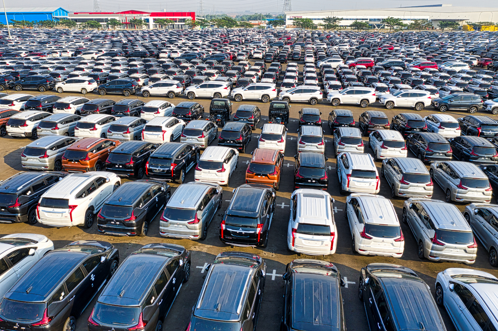 A large parking lot filled with numerous parked cars, with a shopping mall in the background.
