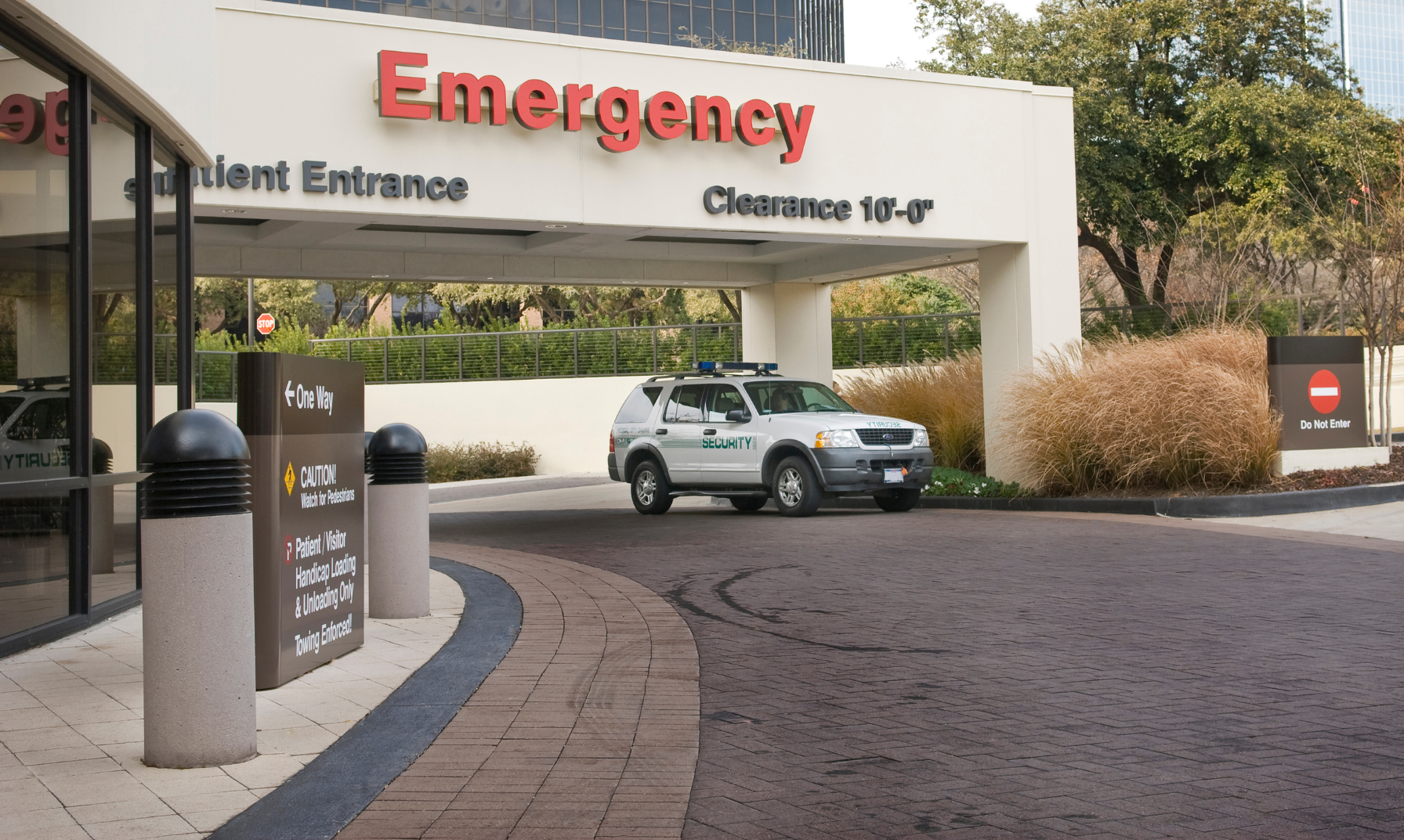 Entrance to hospital emergency department with a security vehicle parked nearby, signs indicating patient entrance, no entry, and directions to one way, with trees and modern buildings in the background.