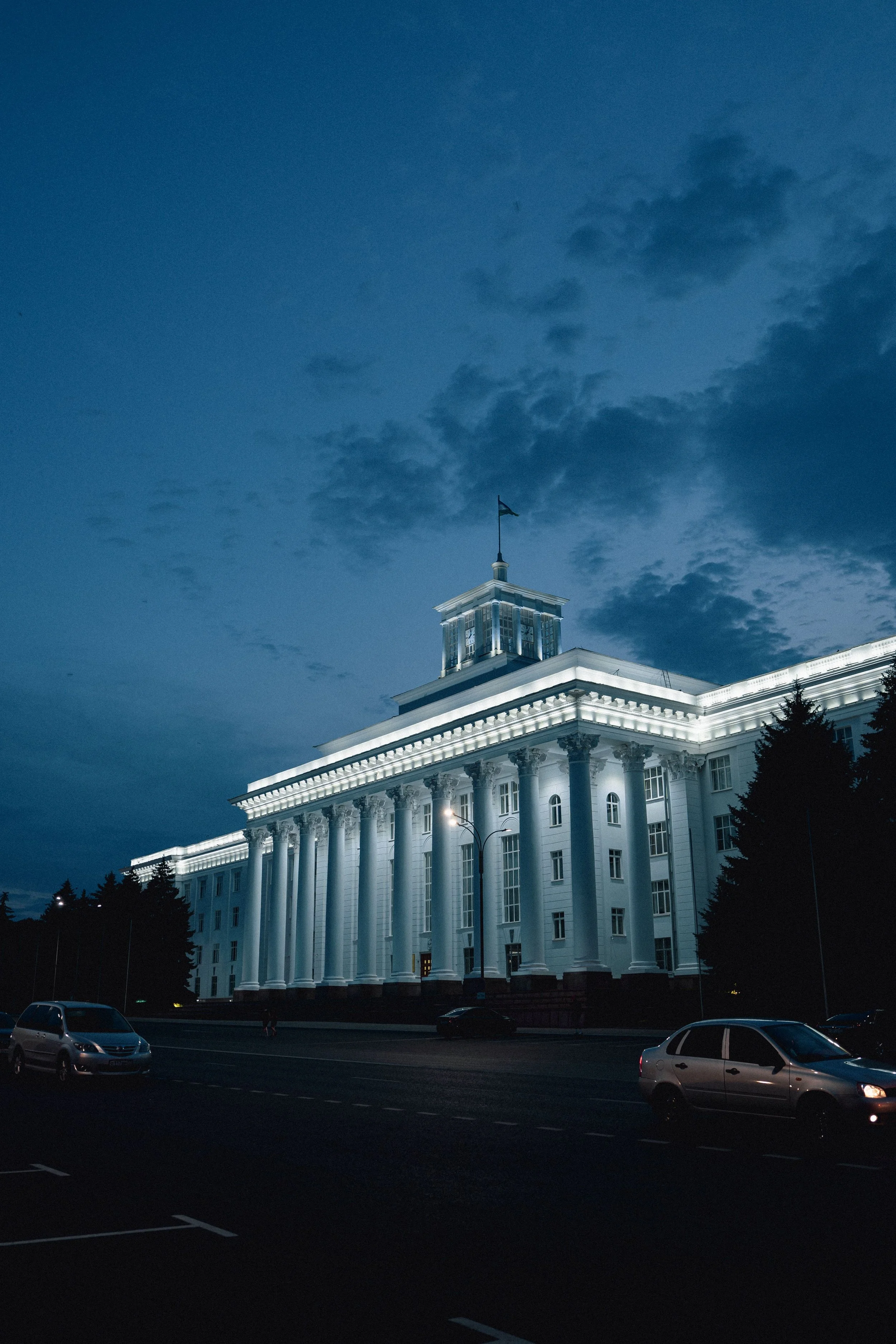 Nighttime view of a large, classic white government building with tall columns and a clock tower, illuminated by exterior lights, with a parking lot and cars in the foreground and dark cloudy sky above.