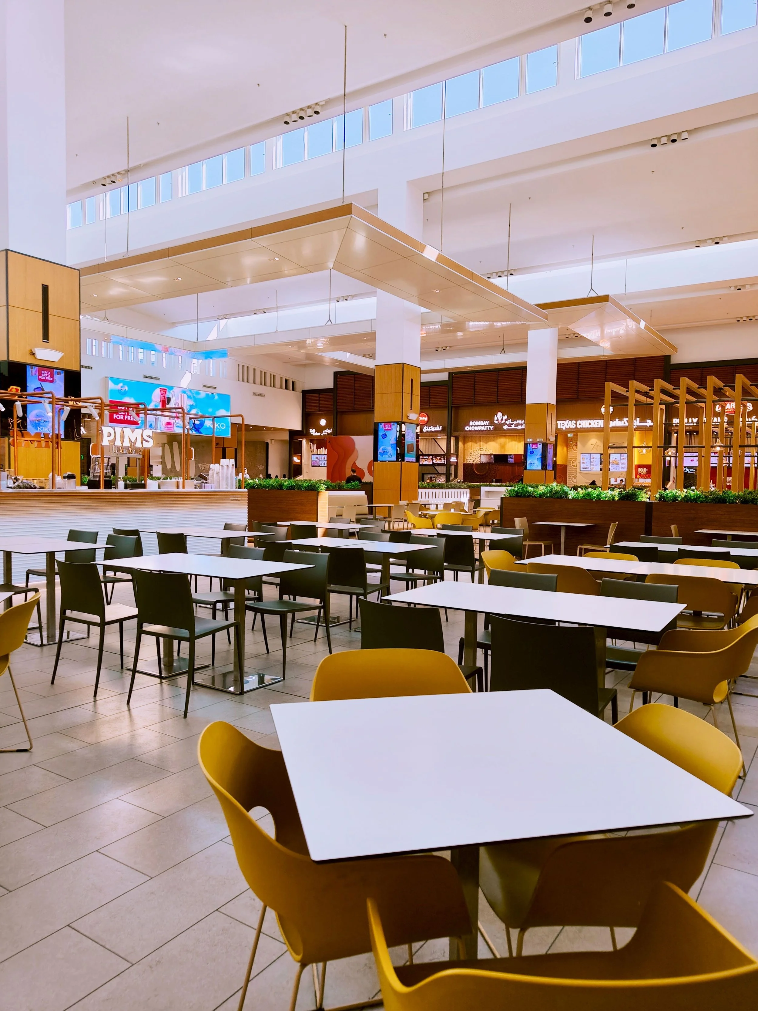 Empty food court area with white tables, yellow and black chairs, and a background of restaurant storefronts in a mall.