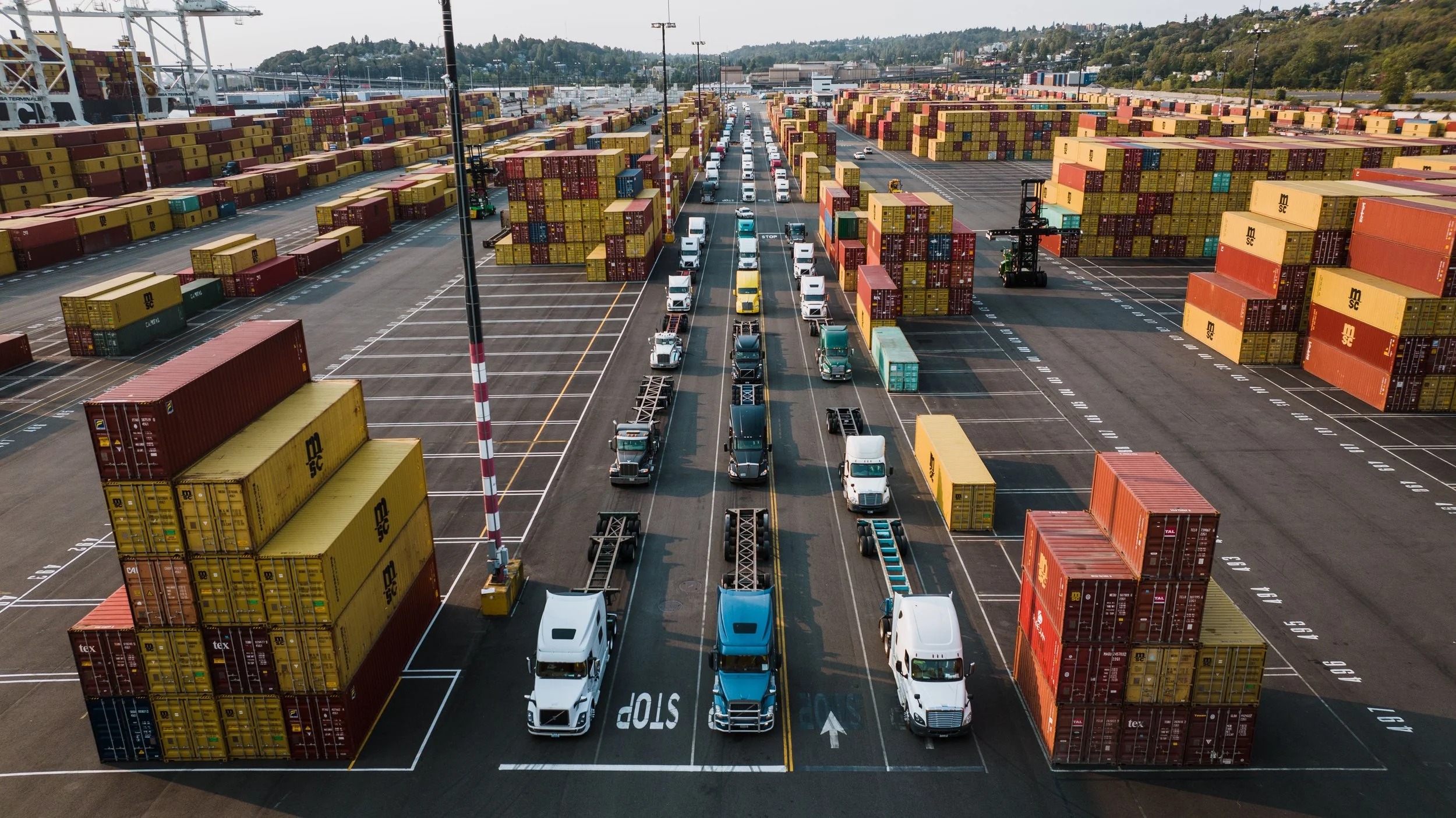 A shipping container yard with rows of stacked containers, trucks lined up for loading or unloading, and a crane in the background.