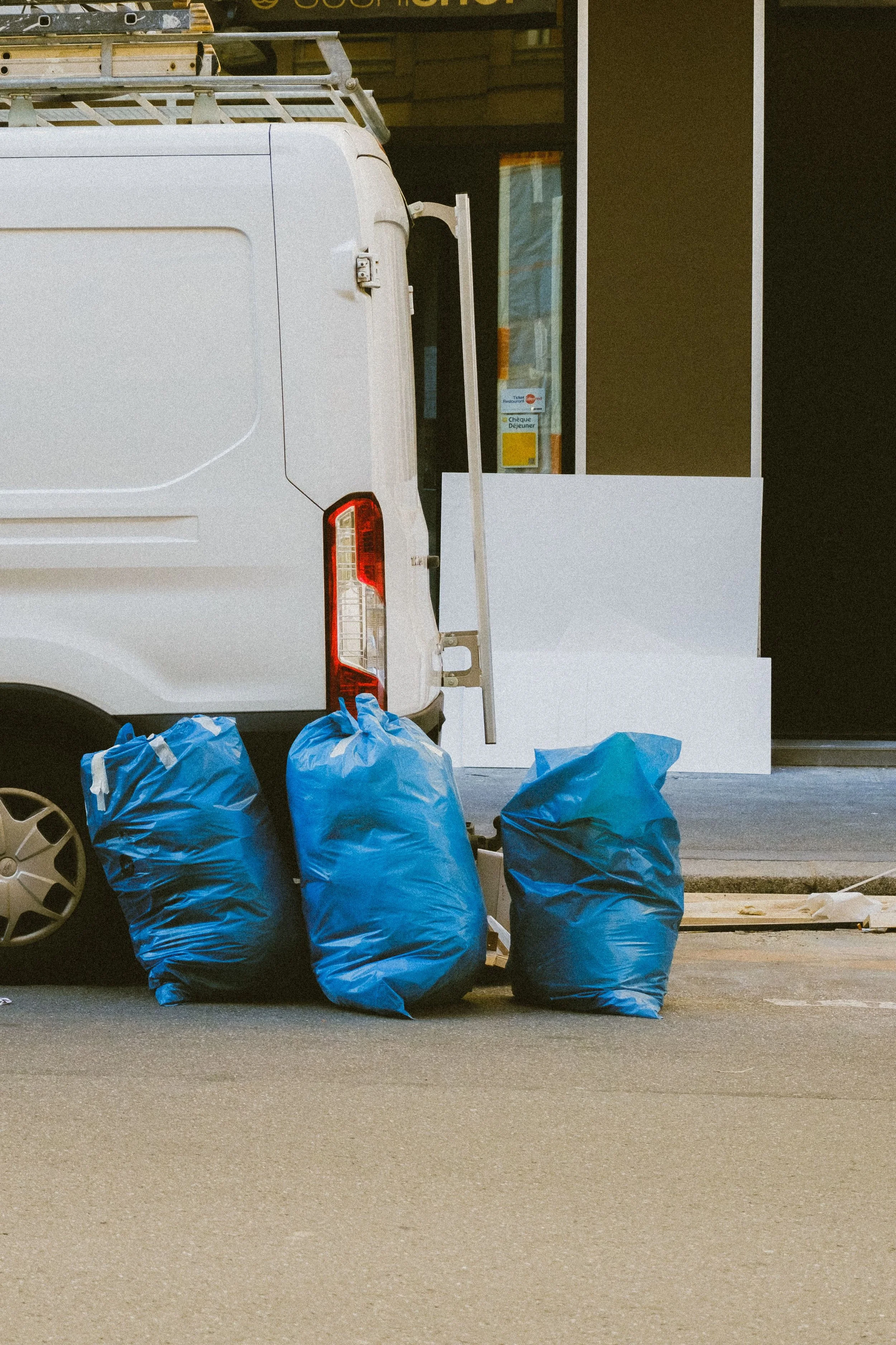 A white delivery van with several filled blue garbage bags in front, parked outside a building with a doorway and window.