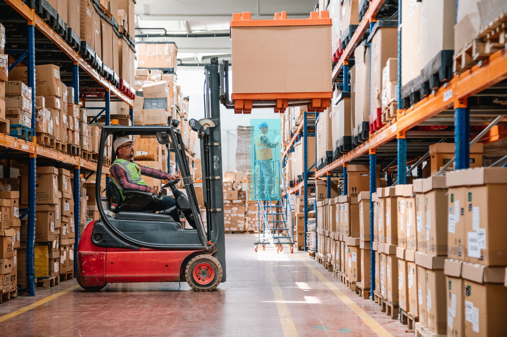 A warehouse worker is operating a forklift to move a large box on a rack. Two workers are in the background, one on a ladder and the other standing nearby, both wearing safety vests and helmets.