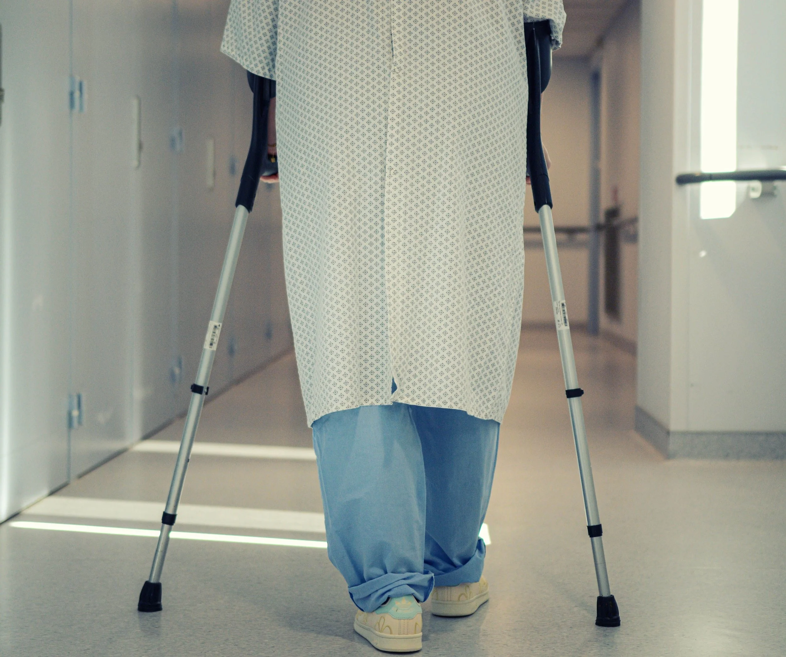 Person in hospital gown walking with crutches in a hospital hallway.