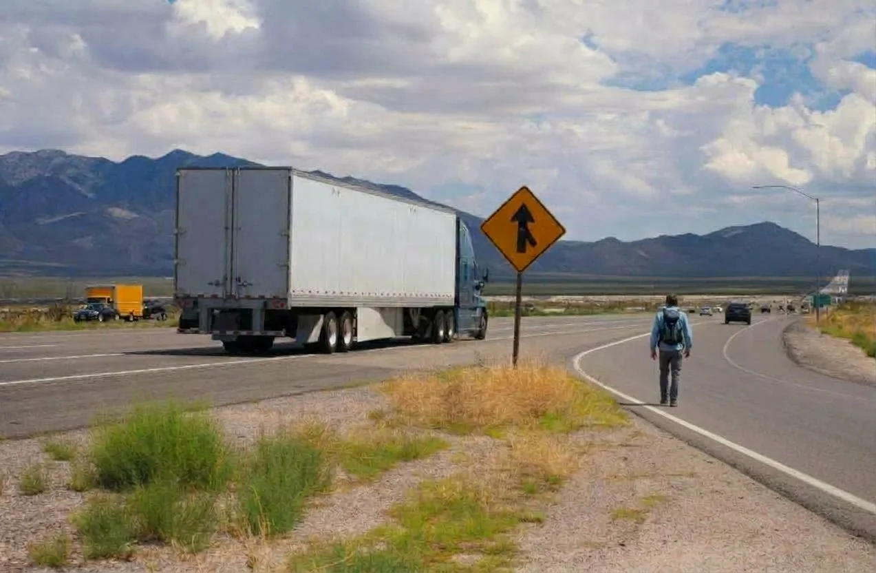 A person walking on the shoulder of a highway next to a large truck, with a mountain range and cloudy sky in the background. There is a yellow road sign indicating merging traffic.