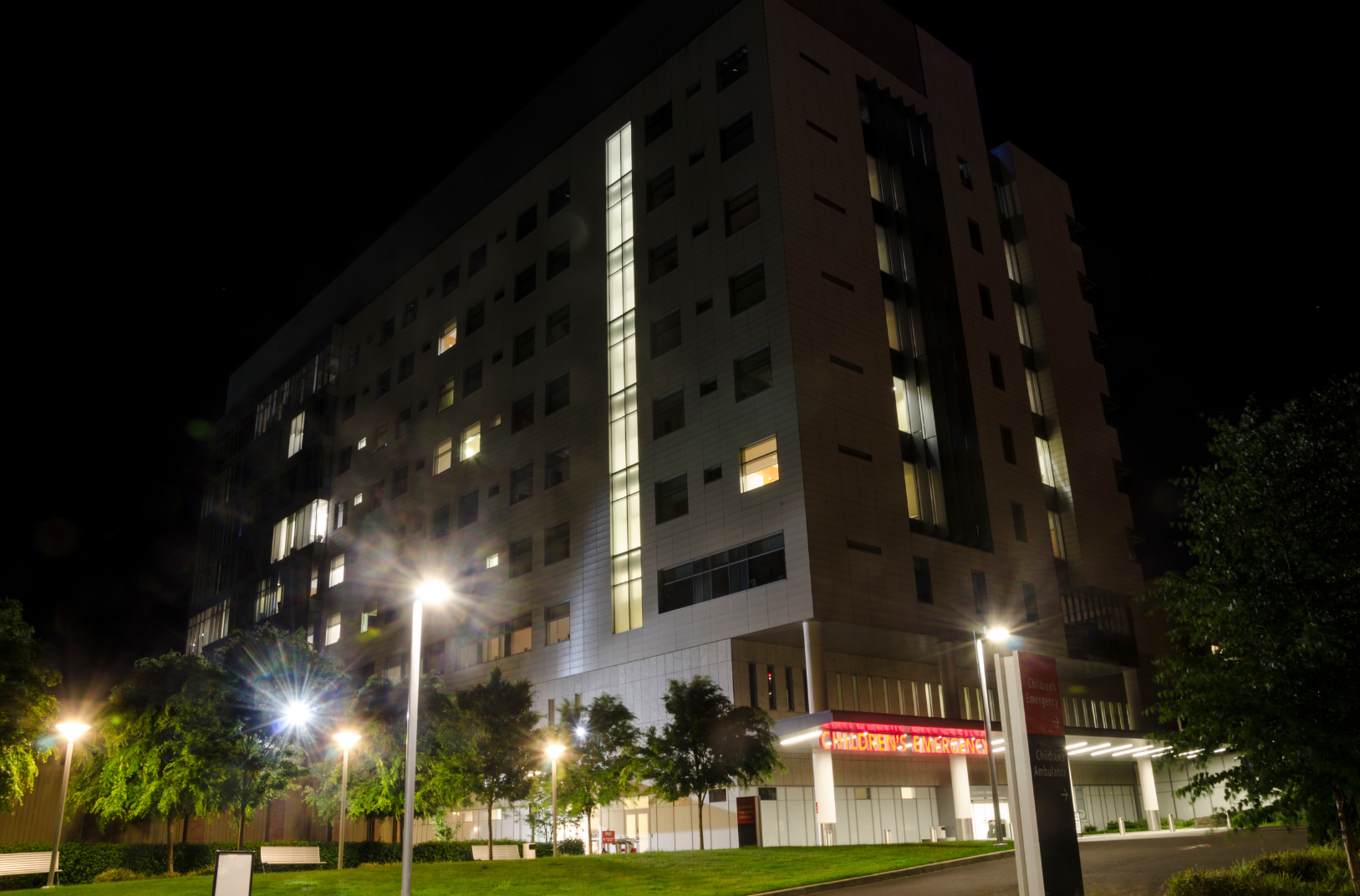 Modern multi-story hospital building at night, illuminated windows, trees and streetlights in foreground, with a sign indicating children's emergency area.