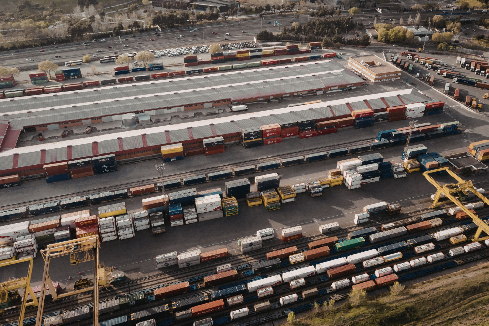An aerial view of a train yard with numerous parked cargo containers and freight vehicles, with a highway in the background.