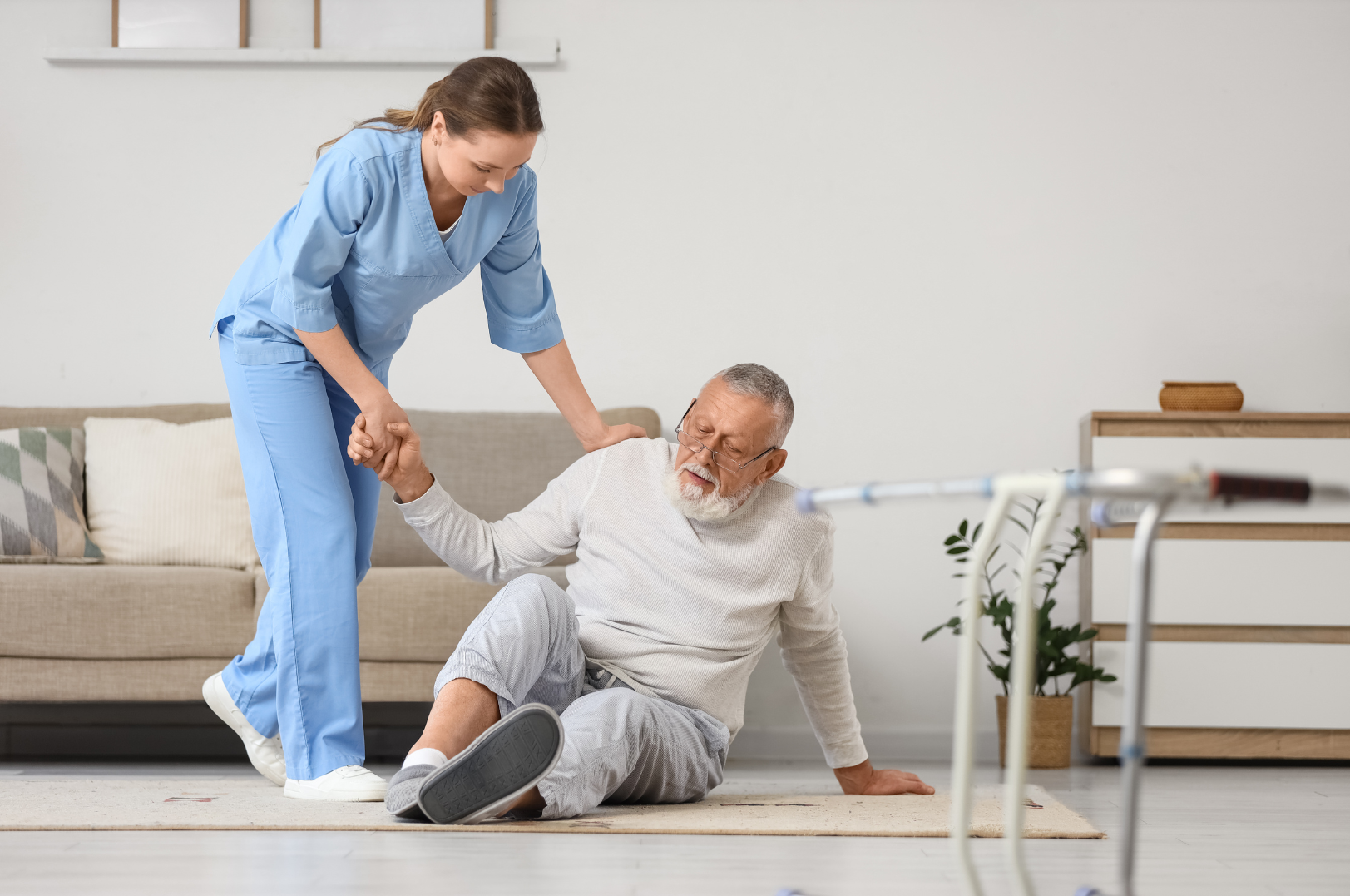 A caregiver helps an elderly man who has fallen on the floor inside a home, with a walker nearby.