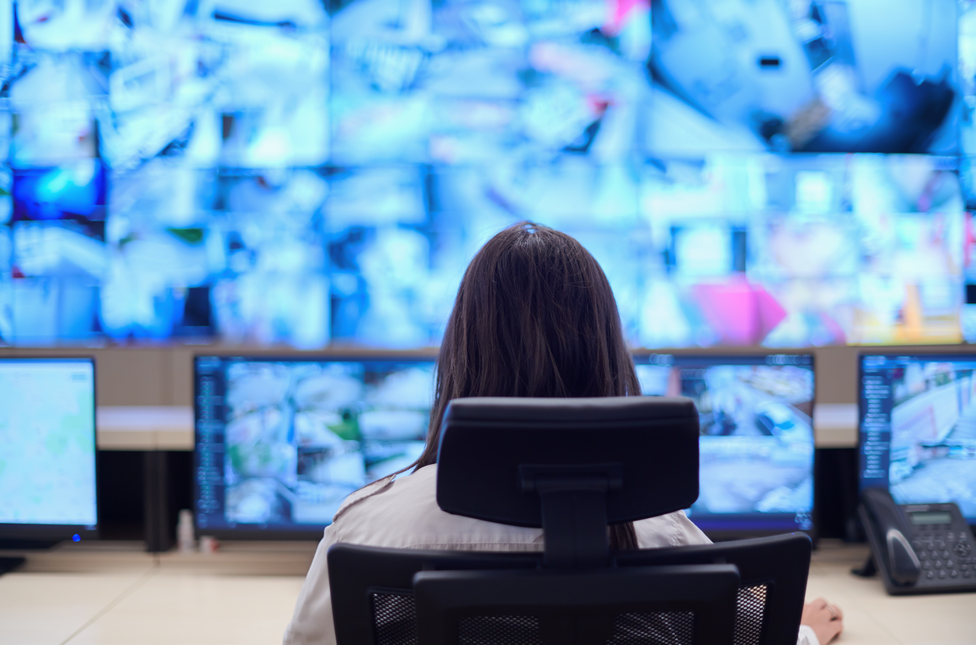 A woman with long brown hair sitting at a desk in a control room or operations center, facing multiple large monitors displaying blurred images with various colors and details.