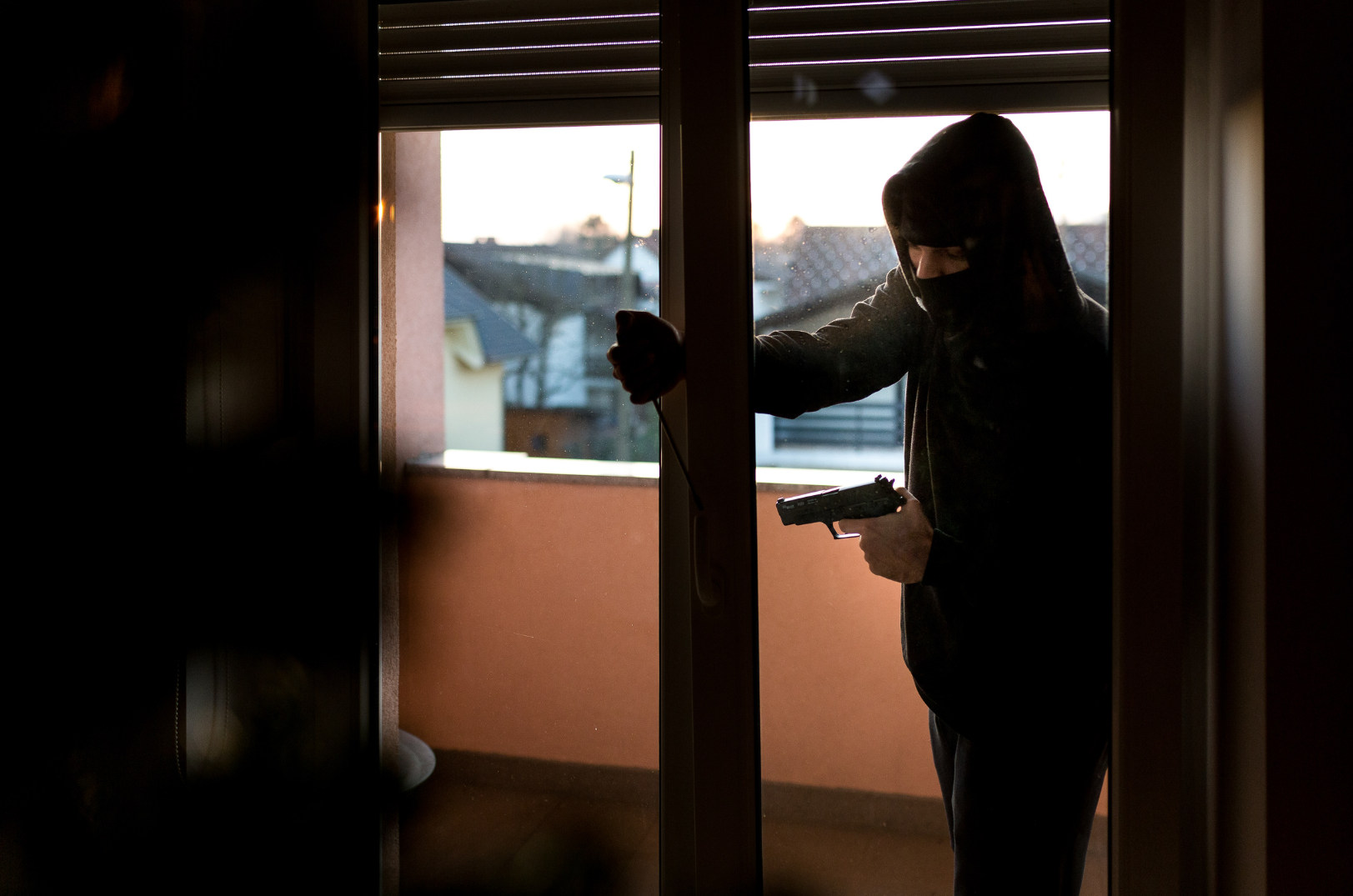 A person wearing a hood and mask standing on a balcony at sunset, entering a sliding glass door holding a handgun and a sledgehammer.