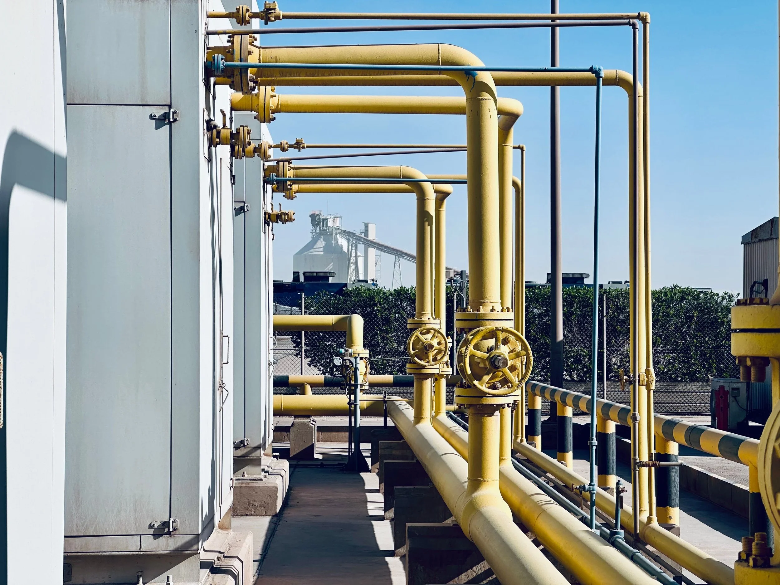 Industrial setting with yellow pipelines and valves, with a white electrical box on the left side, and an industrial plant with large structures in the background under a blue sky.