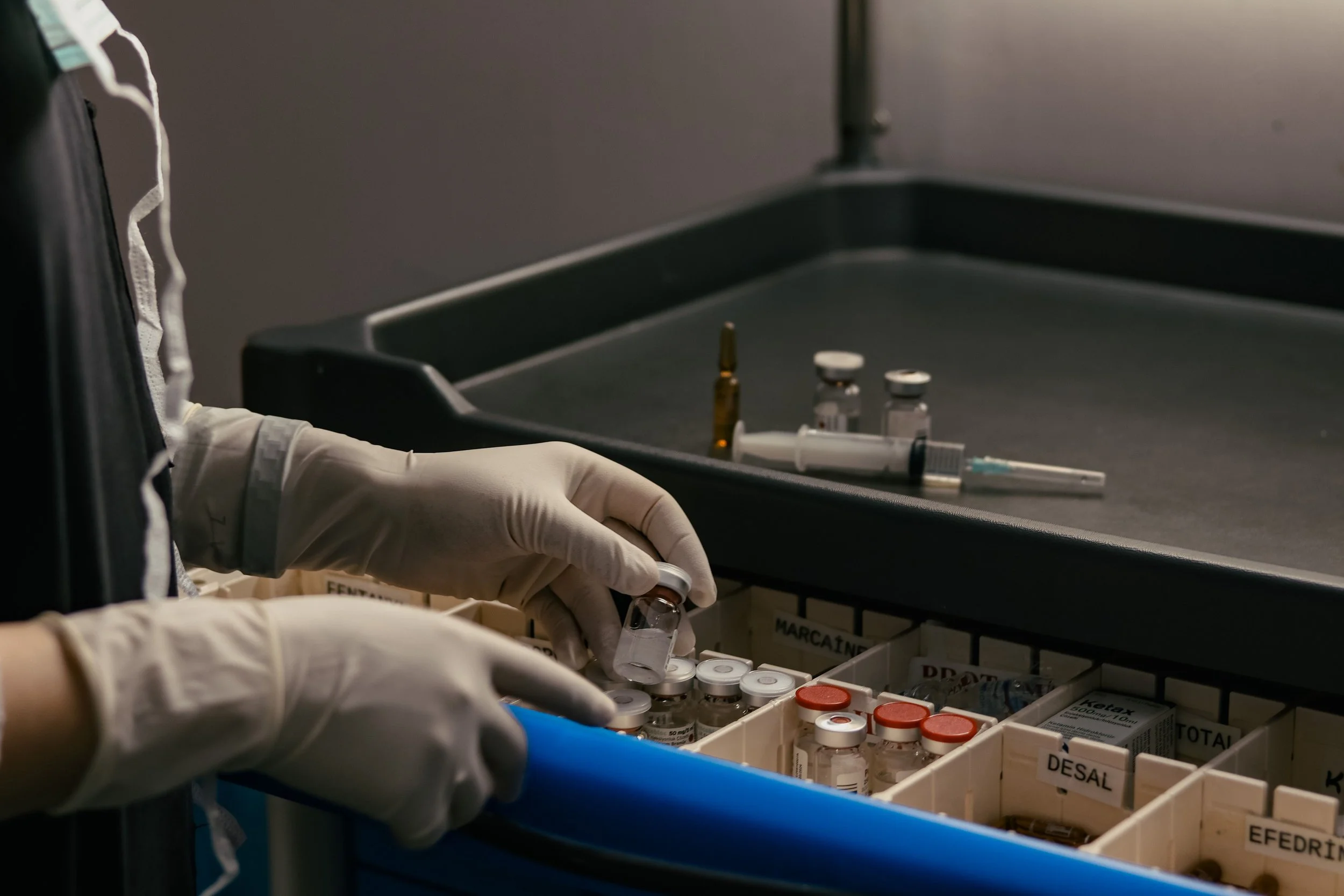 A healthcare worker wearing white gloves handling small vials of medication or vaccine in a medical setting, with a black tray and labeled storage compartments in the background.