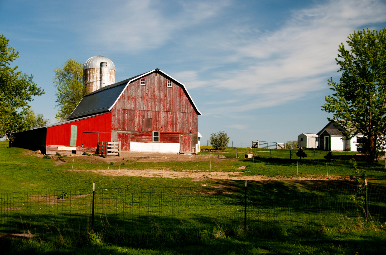 A red barn with a silo in a green field under a blue sky with scattered clouds.