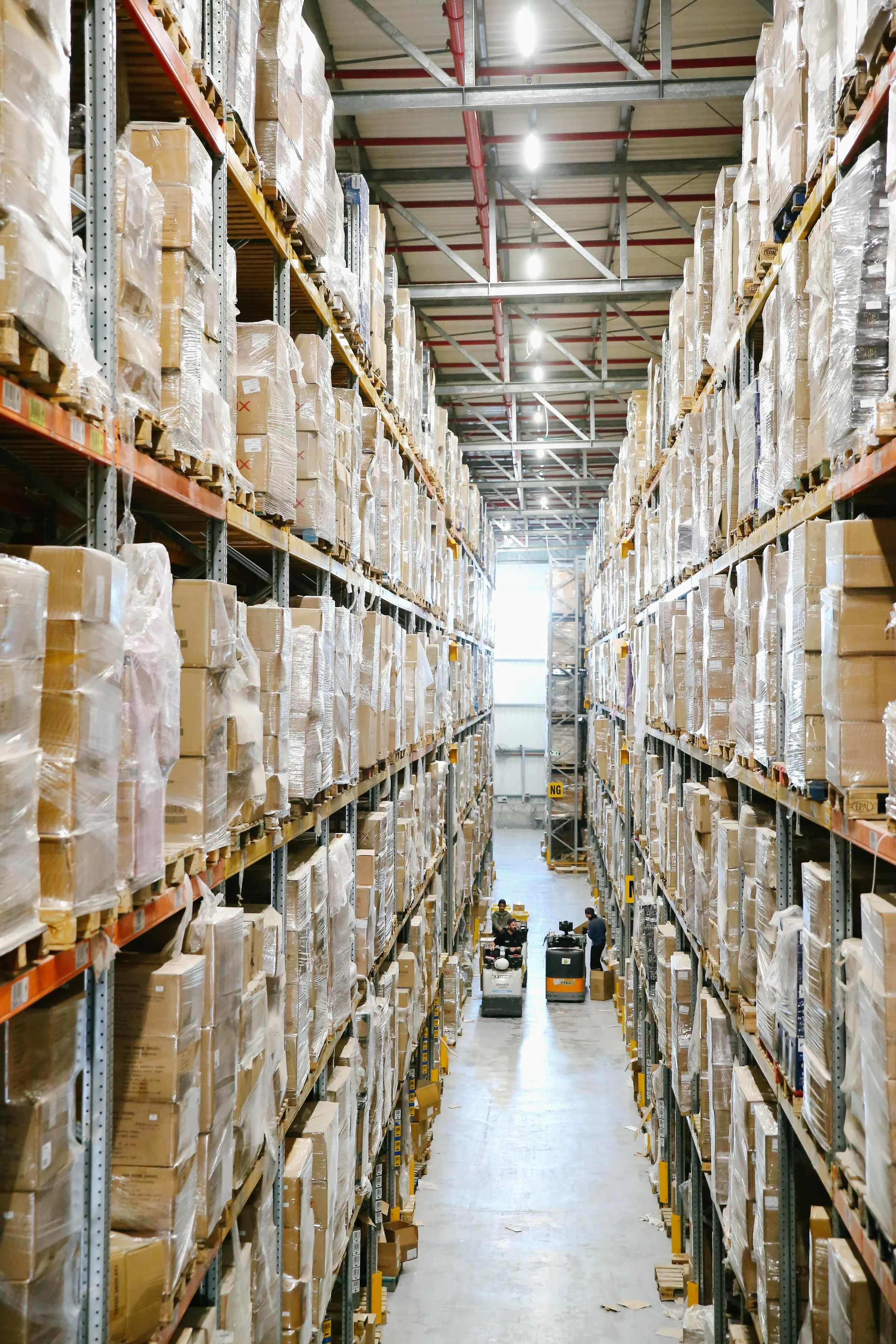 An aisle in a warehouse with tall shelves filled with boxed goods wrapped in plastic. There are workers operating forklifts and sorting items.