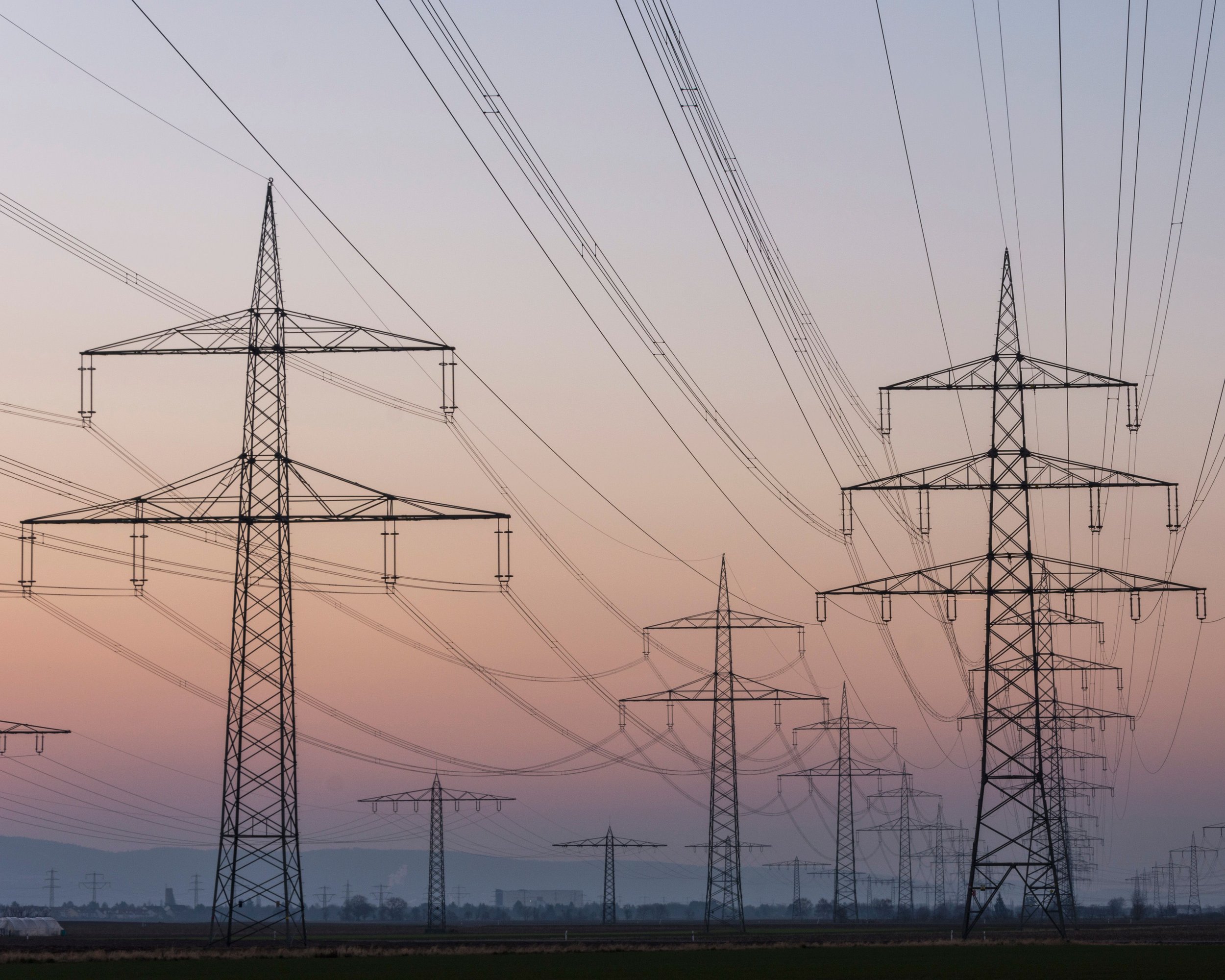 Multiple electrical transmission towers and power lines against a sunset or dawn sky with a gradient of light colors.