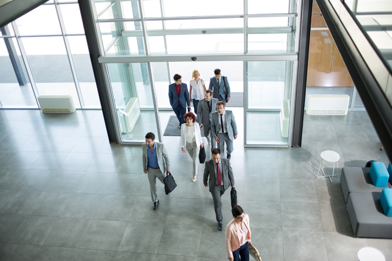 A group of businesspeople walking out of an office building into a lobby area with glass walls and modern furniture.