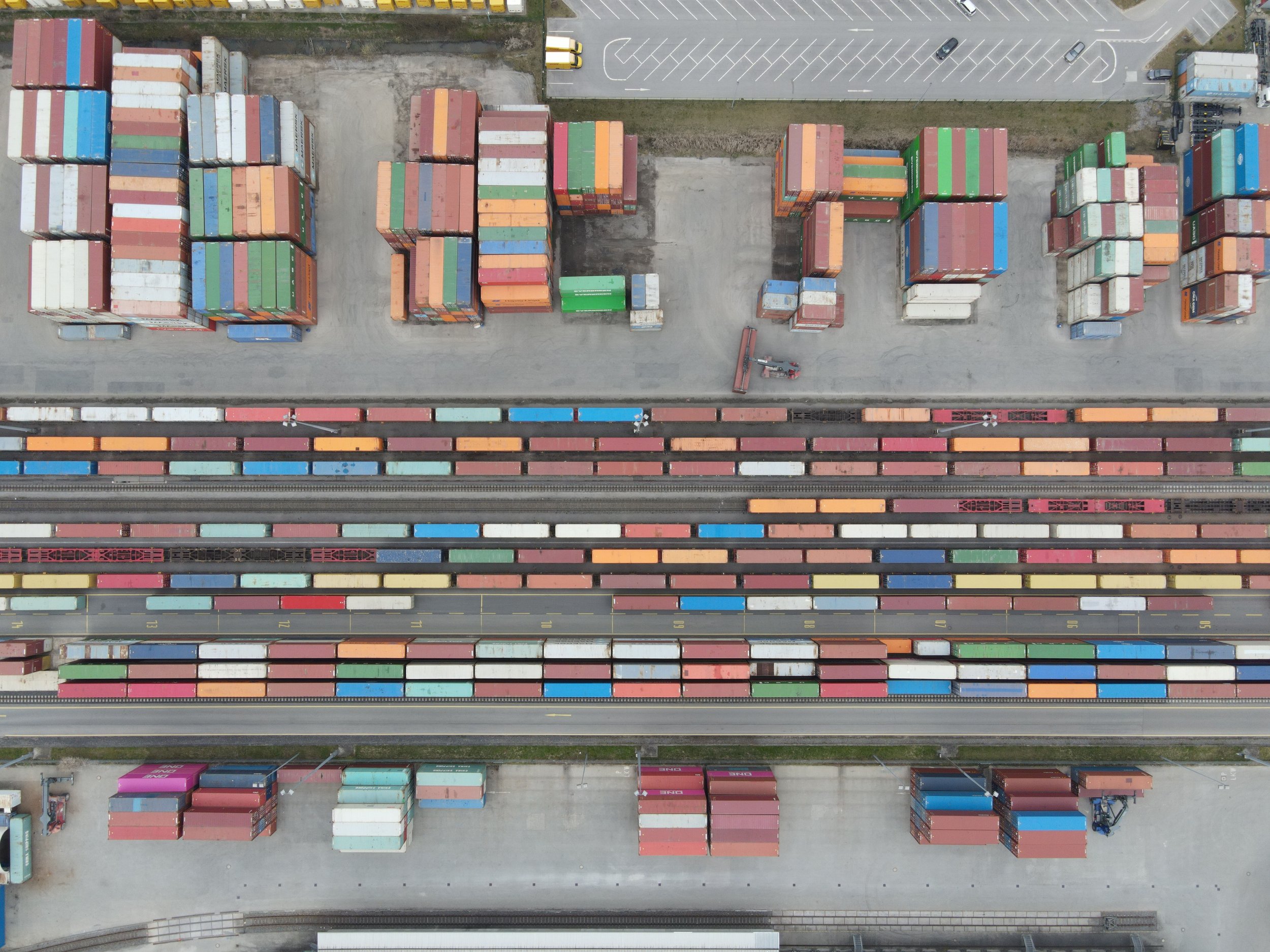 An aerial view of a shipping yard with numerous stacked containers in various colors, including red, green, orange, blue, and white, aligned in rows and separated by roads.