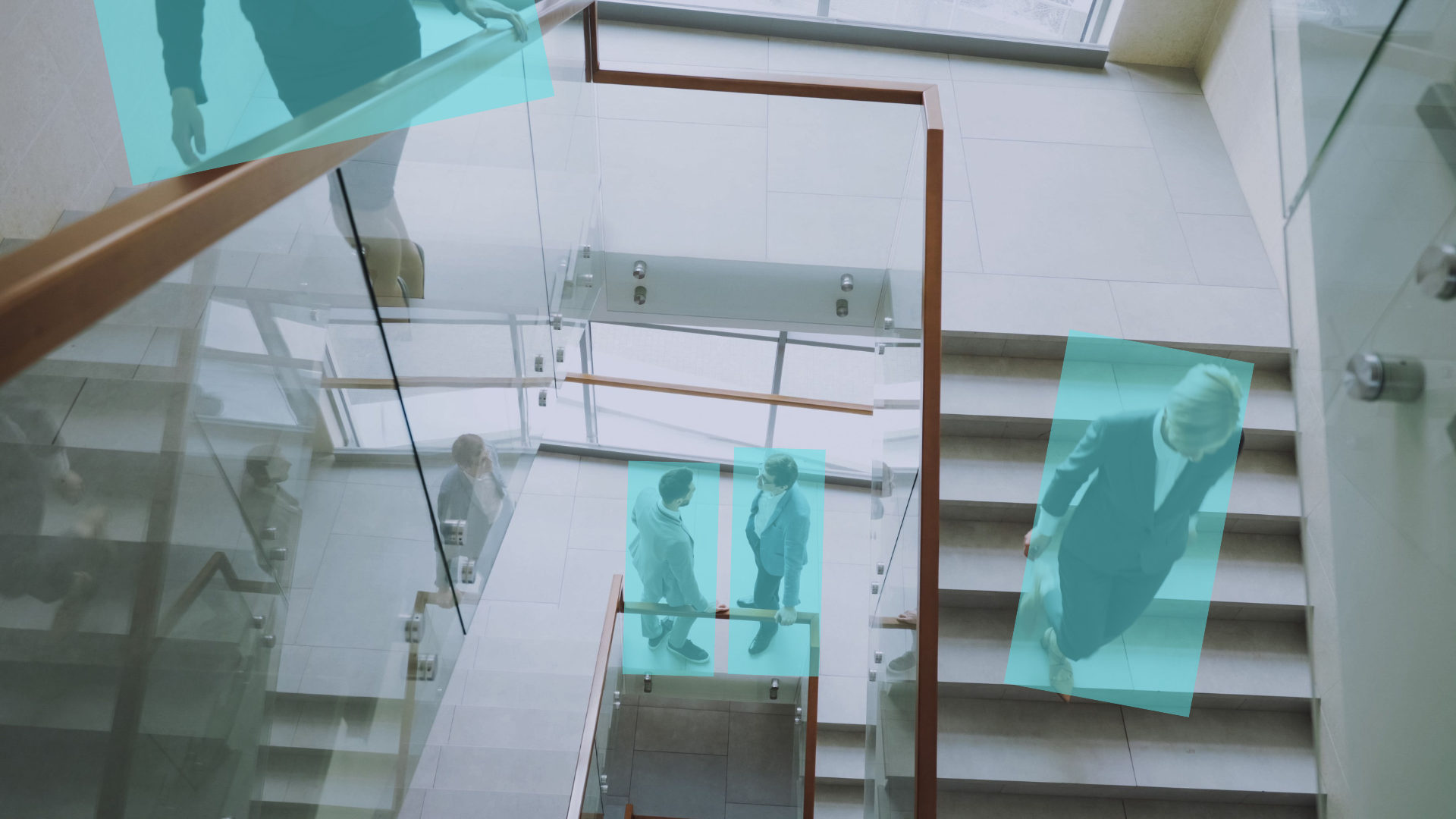 Interior view of a modern, multi-story office building stairwell taken from the top looking down. Glass walls & doors reflect the surroundings. Several people are walking through the space, highlighted in blue to indicate the people counting feature.