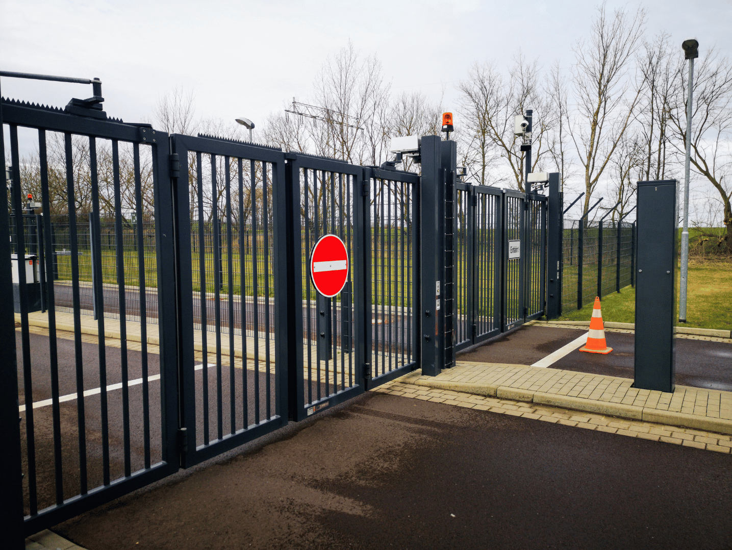 Electric gate with a red 'no entry' sign, safety cones, and security cameras, set outdoors with trees and overcast sky in the background.