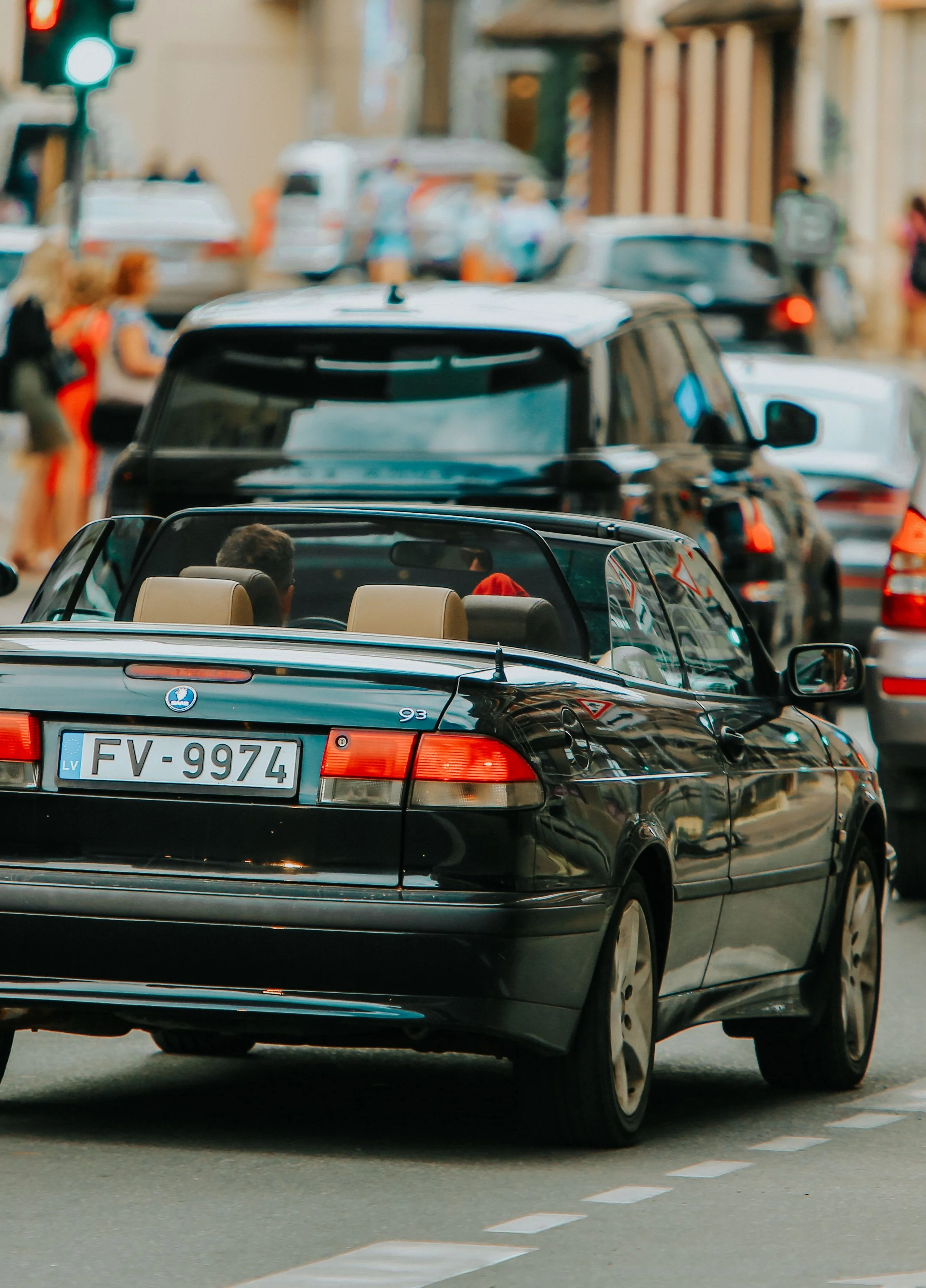 Several cars driving on a city street with pedestrians walking nearby and a traffic light showing green.