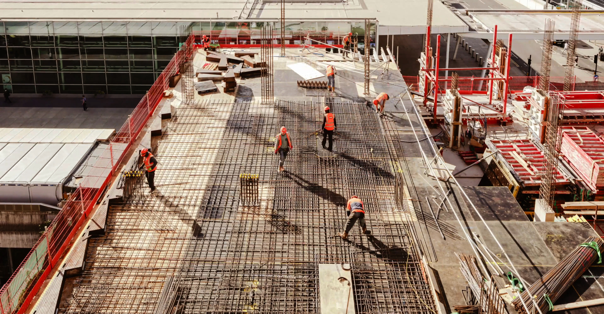 Construction workers in safety vests and helmets working on steel rebar framework on a high-rise building under construction, with a cityscape in the background.
