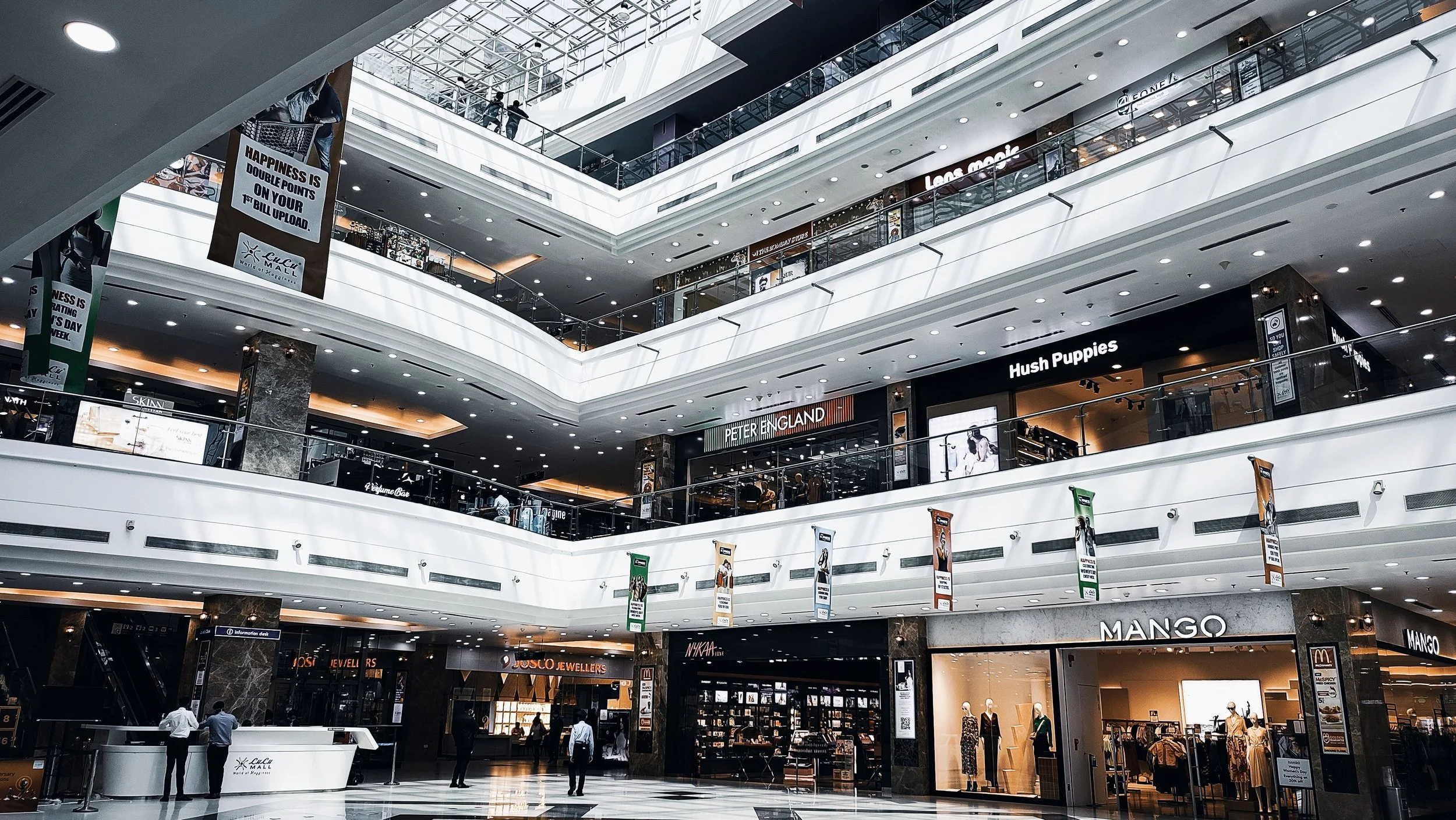 Interior view of a multi-level shopping mall with stores like Hush Puppies, Mango, and Peter England, escalators, banners, and shoppers walking inside.