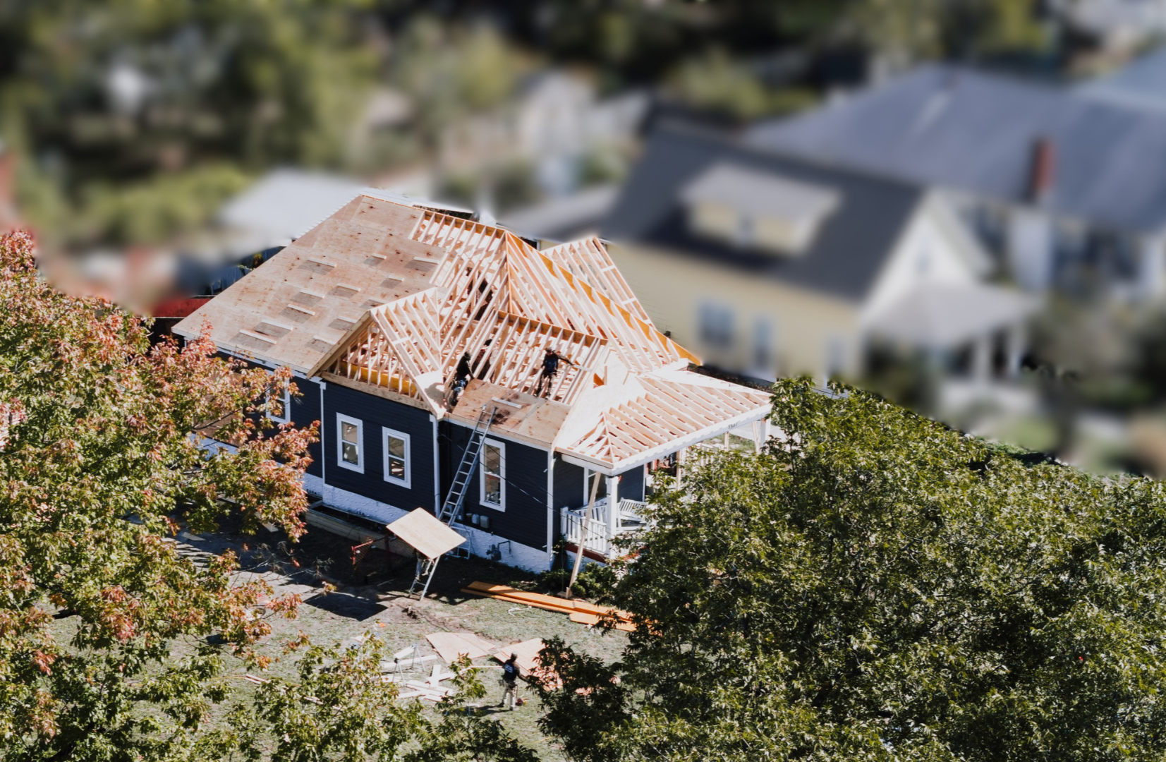 A house under construction with a wooden roof frame, surrounded by green trees and other houses in the neighborhood, taken from an aerial view.