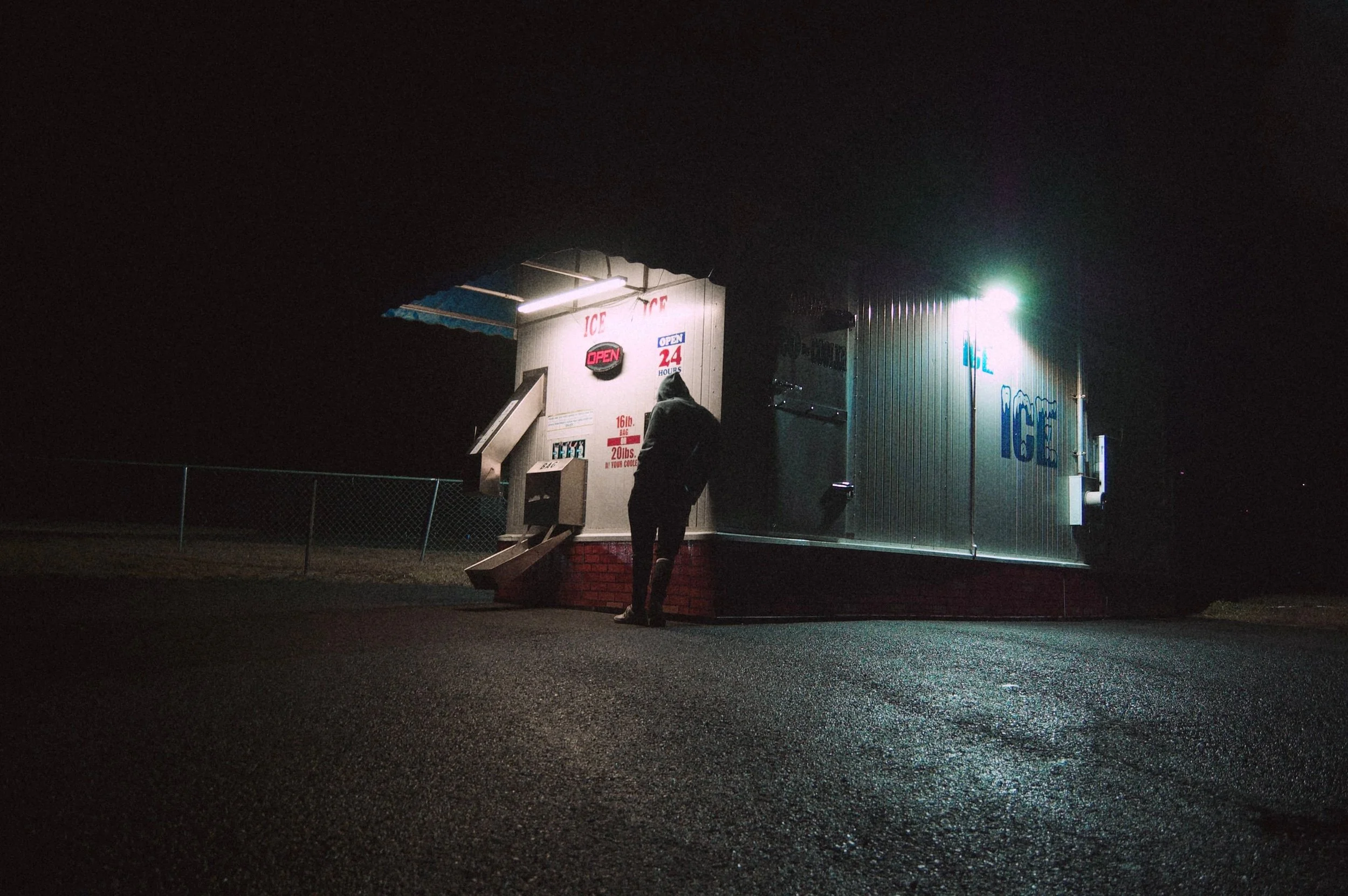 Nighttime scene of a small ice stand with a person standing outside, illuminated by bright lights, against a dark sky.