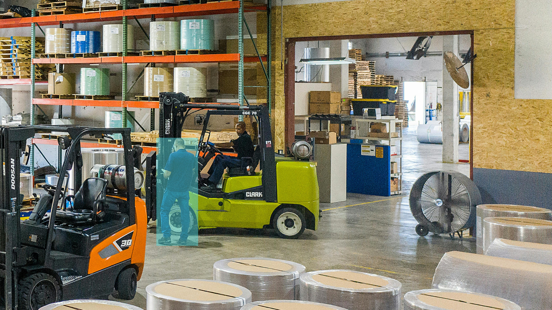 Warehouse interior with shelves stocked with large rolls of material, forklifts and workers, some operating machinery, and large industrial fans.