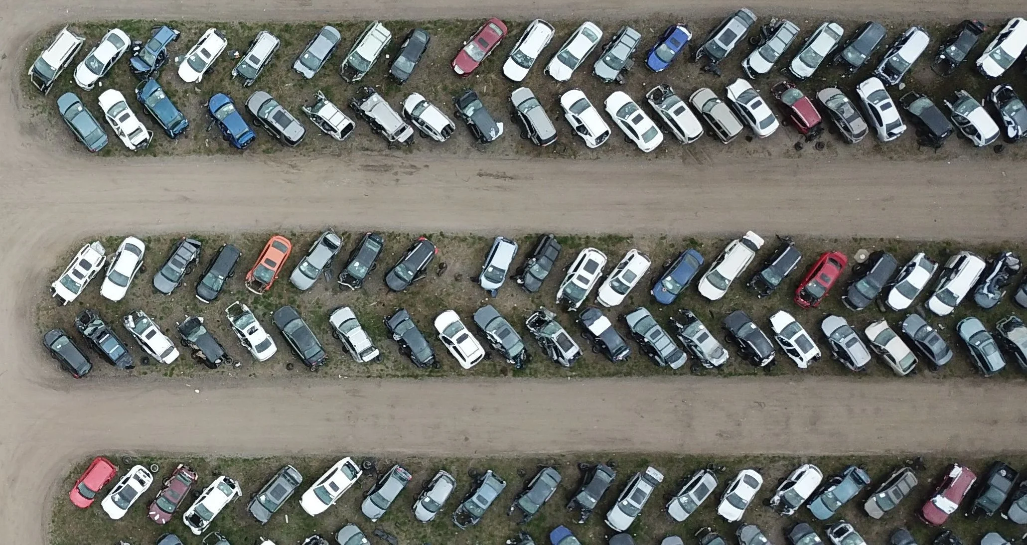 Aerial view of multiple parked cars in a dirt lot, separated by grassy strips, with a dirt road running parallel to the parking rows.