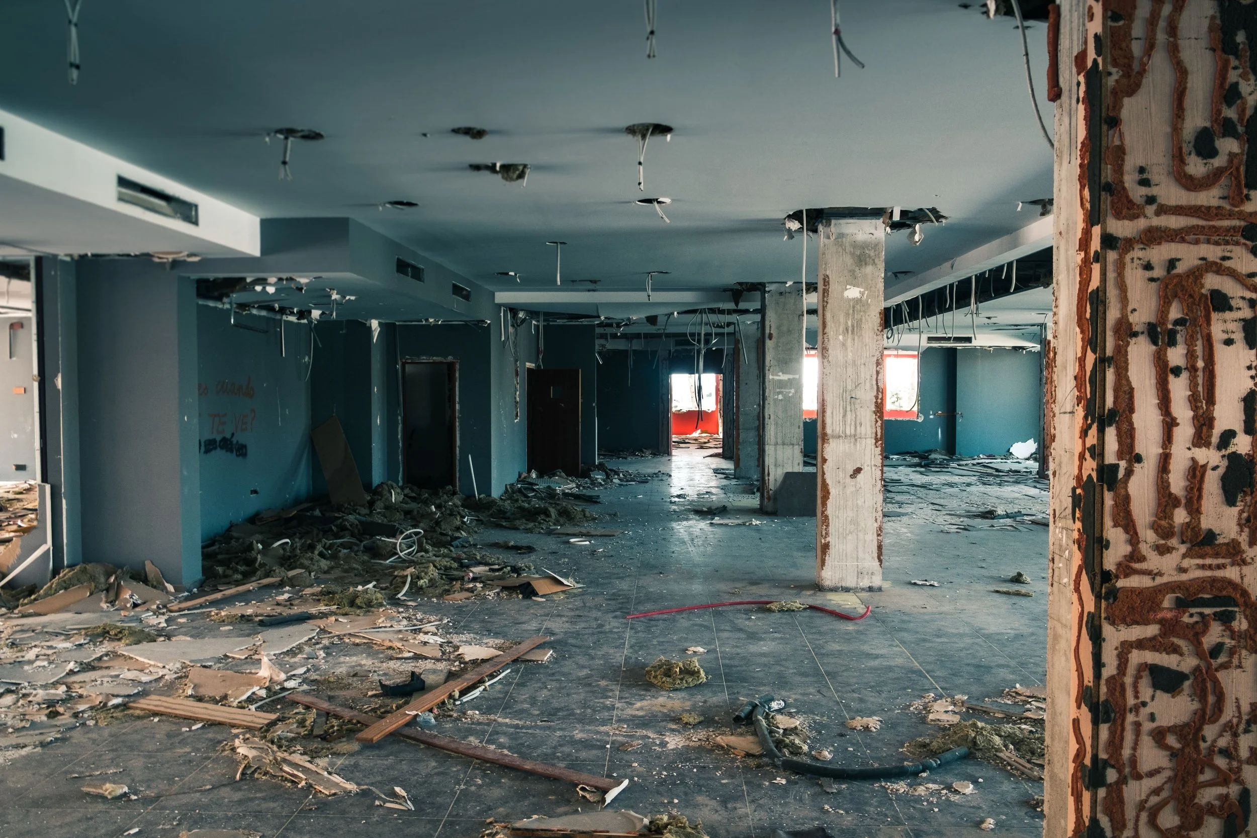 An empty and abandoned interior of a building with damaged ceiling, exposed wiring, debris on the floor, and partially torn walls.