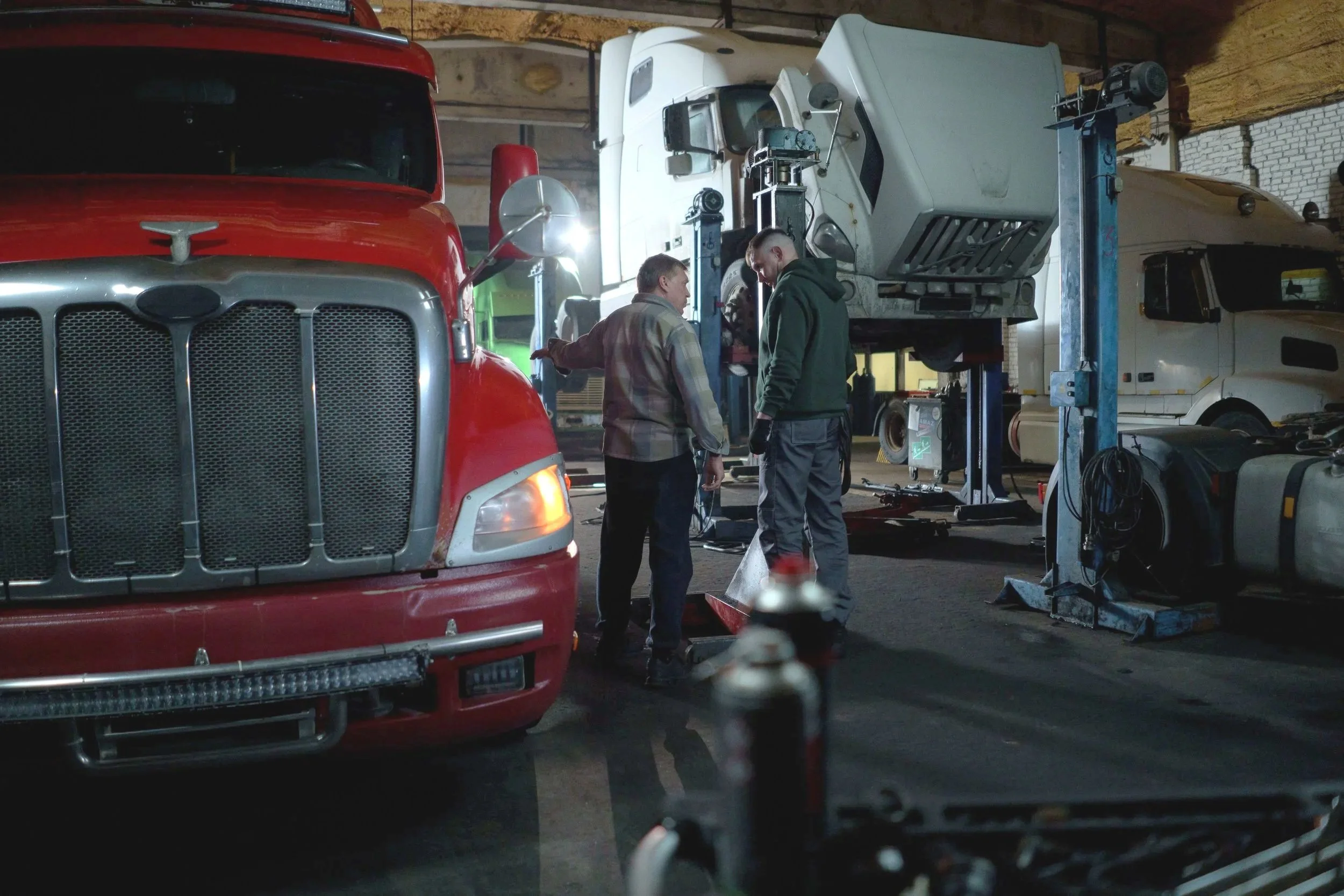 Two men working in a garage or workshop with various semi-trucks and equipment around, including a red semi-truck in the foreground and a white truck on a lift in the background.