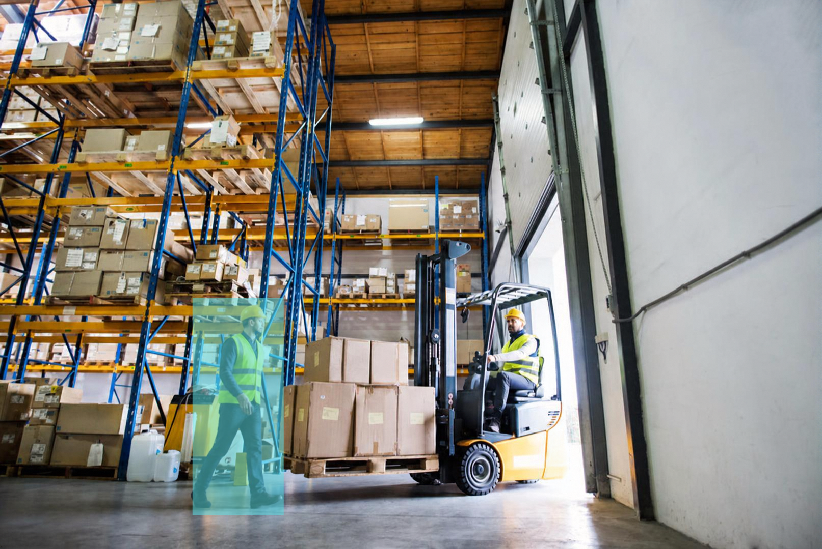 Warehouse with tall shelves filled with boxes, two workers with safety vests and helmets, one operating a forklift and another walking nearby.