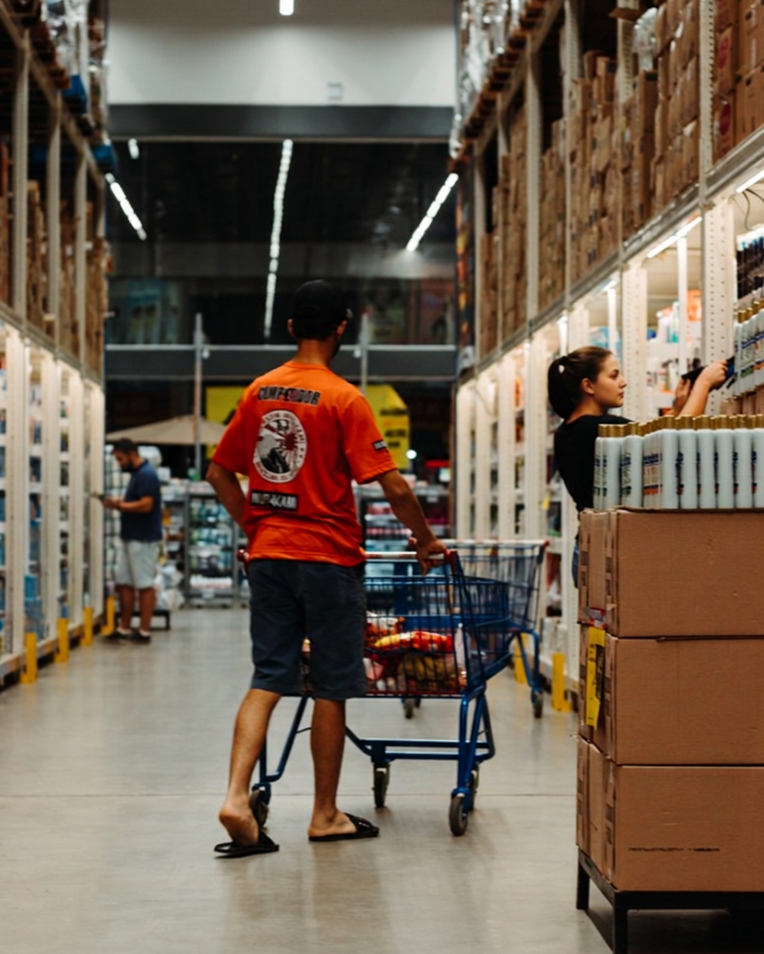 A man with a dark cap, orange t-shirt, and shorts shopping in a store aisle with a shopping cart, and a woman with a ponytail reaching for items on the shelf.