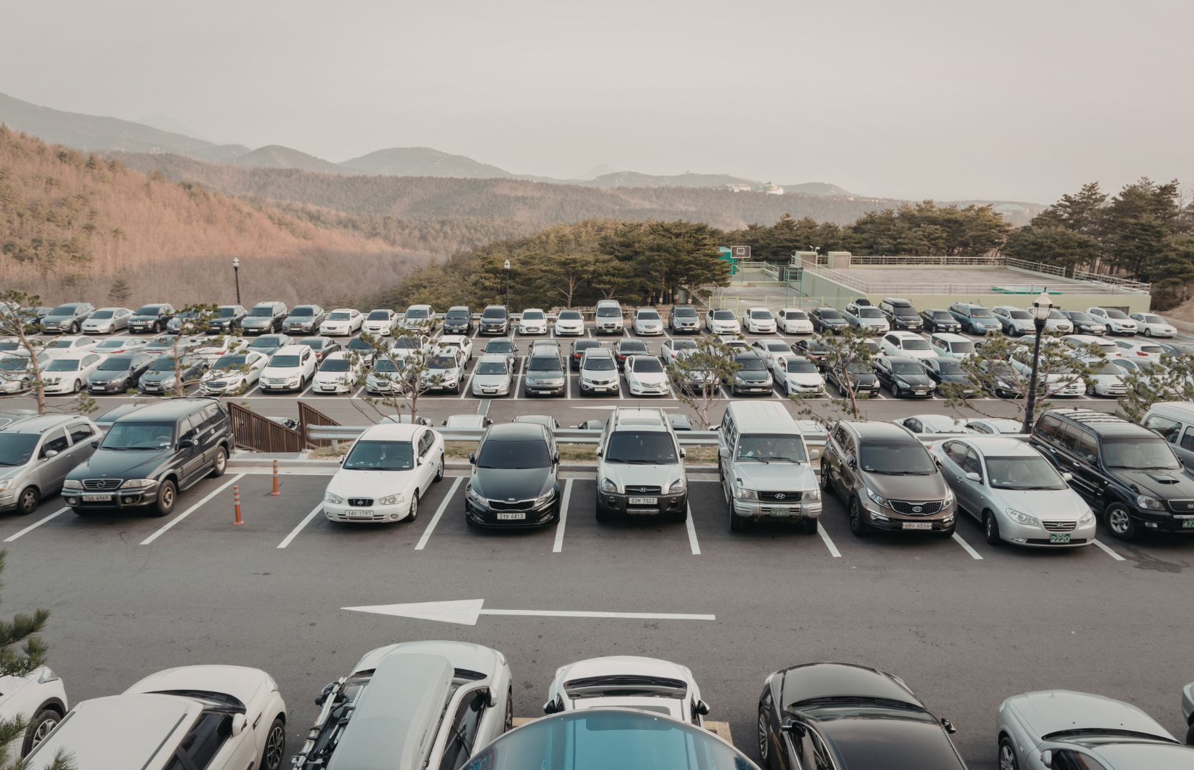 A parking lot filled with various cars, with a mountainous landscape in the background and trees around the area.