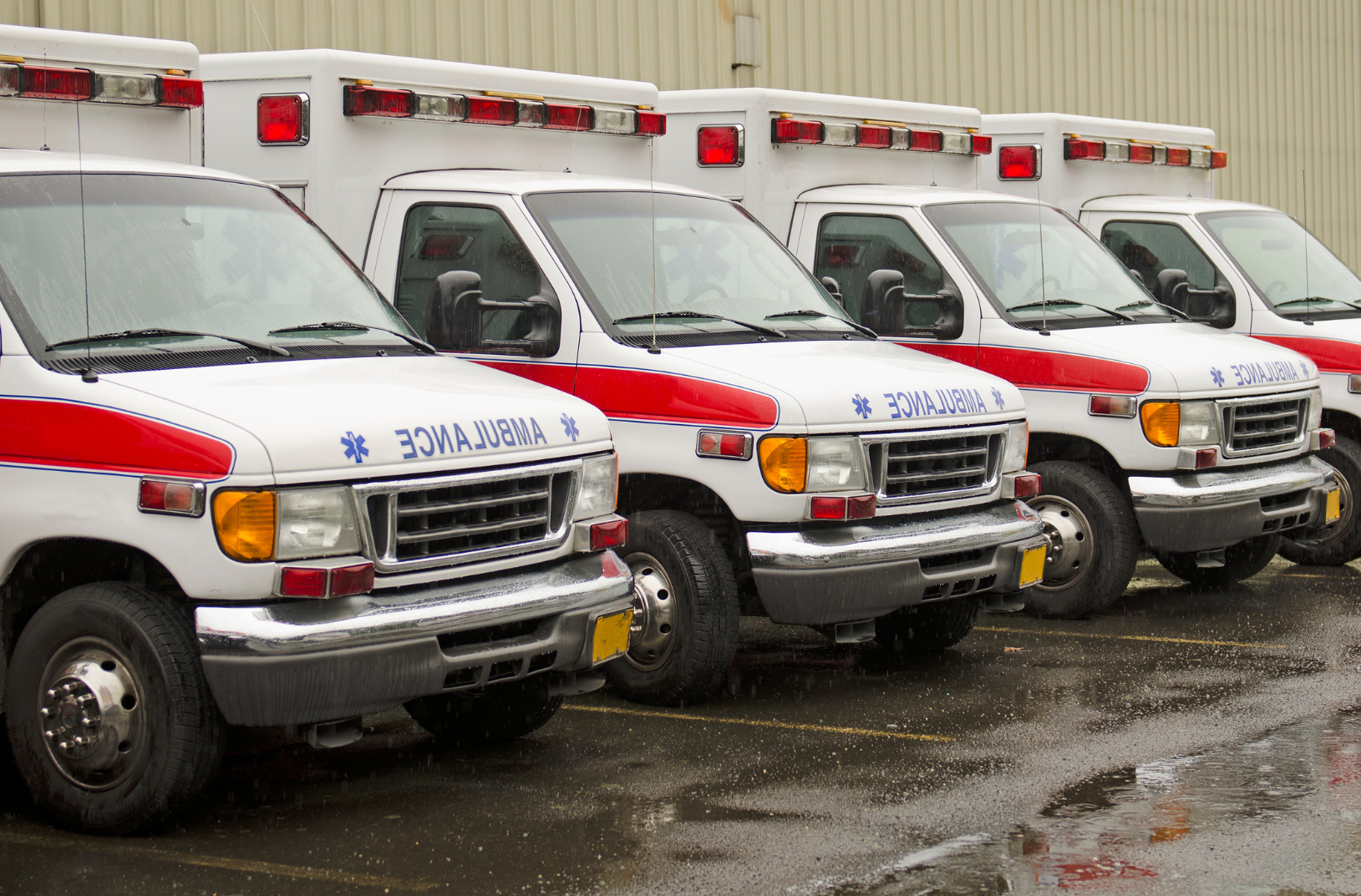 A lineup of four ambulances parked outdoors on a wet surface, with a metal building in the background.