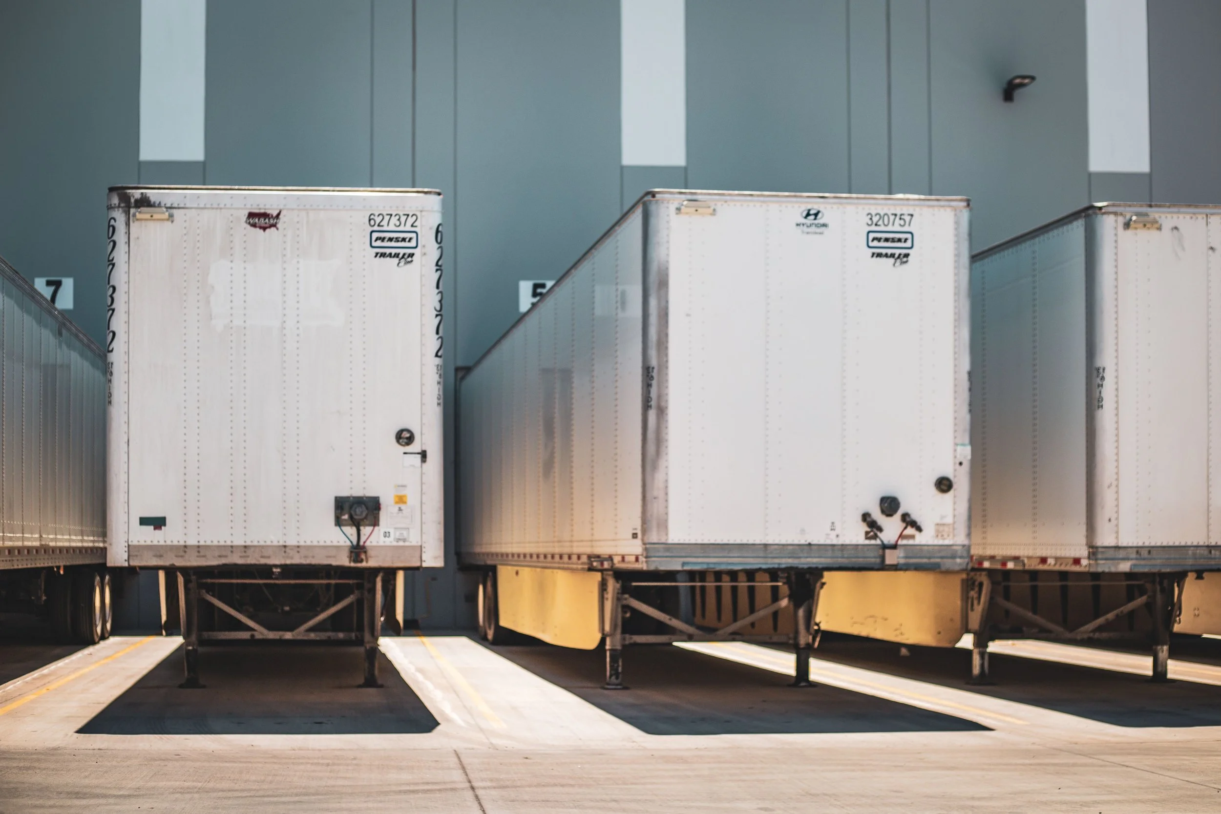 A row of three semi-trailer trucks parked at a loading dock during the middle of the day.