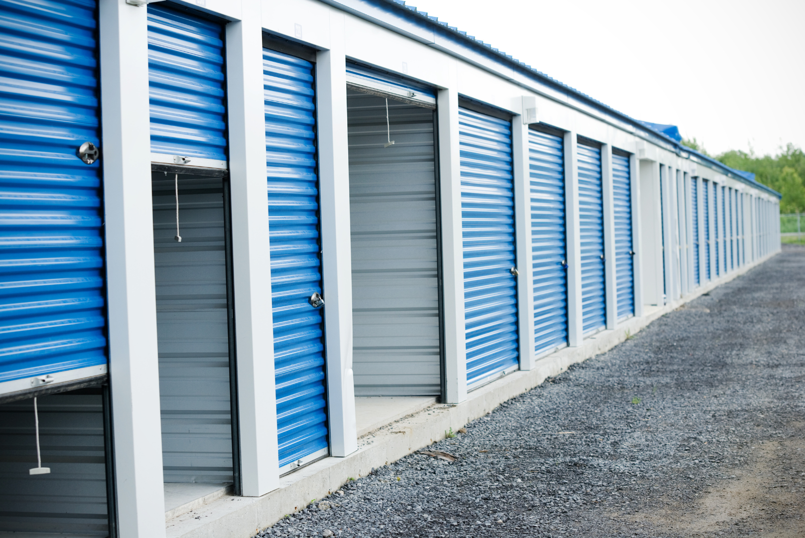 A row of blue storage unit doors, some of which are slightly open, with a gravel pathway in front.
