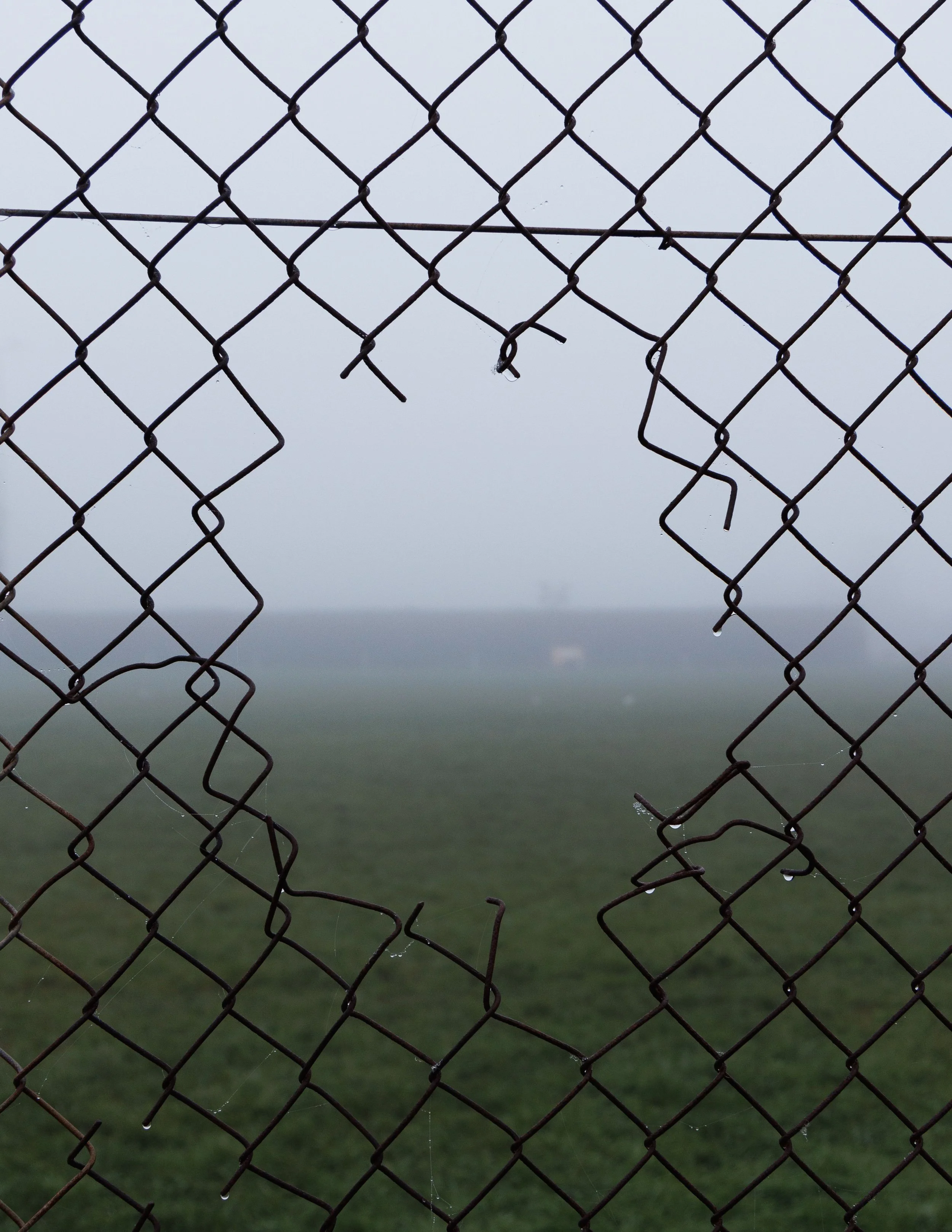 Close-up of a weathered metal chain-link fence with a large hole in it, with the backdrop of foggy, overcast sky and a blurry green landscape.
