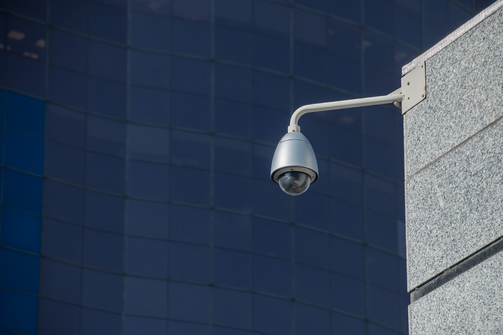 A security camera mounted on a building wall with a background of dark blue reflective glass windows.
