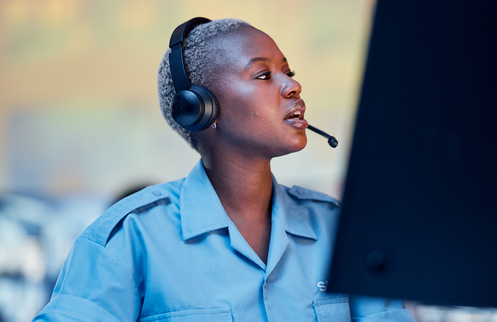 A woman with crew cut gray hair wearing a blue uniform and black headset with microphone working at a computer.