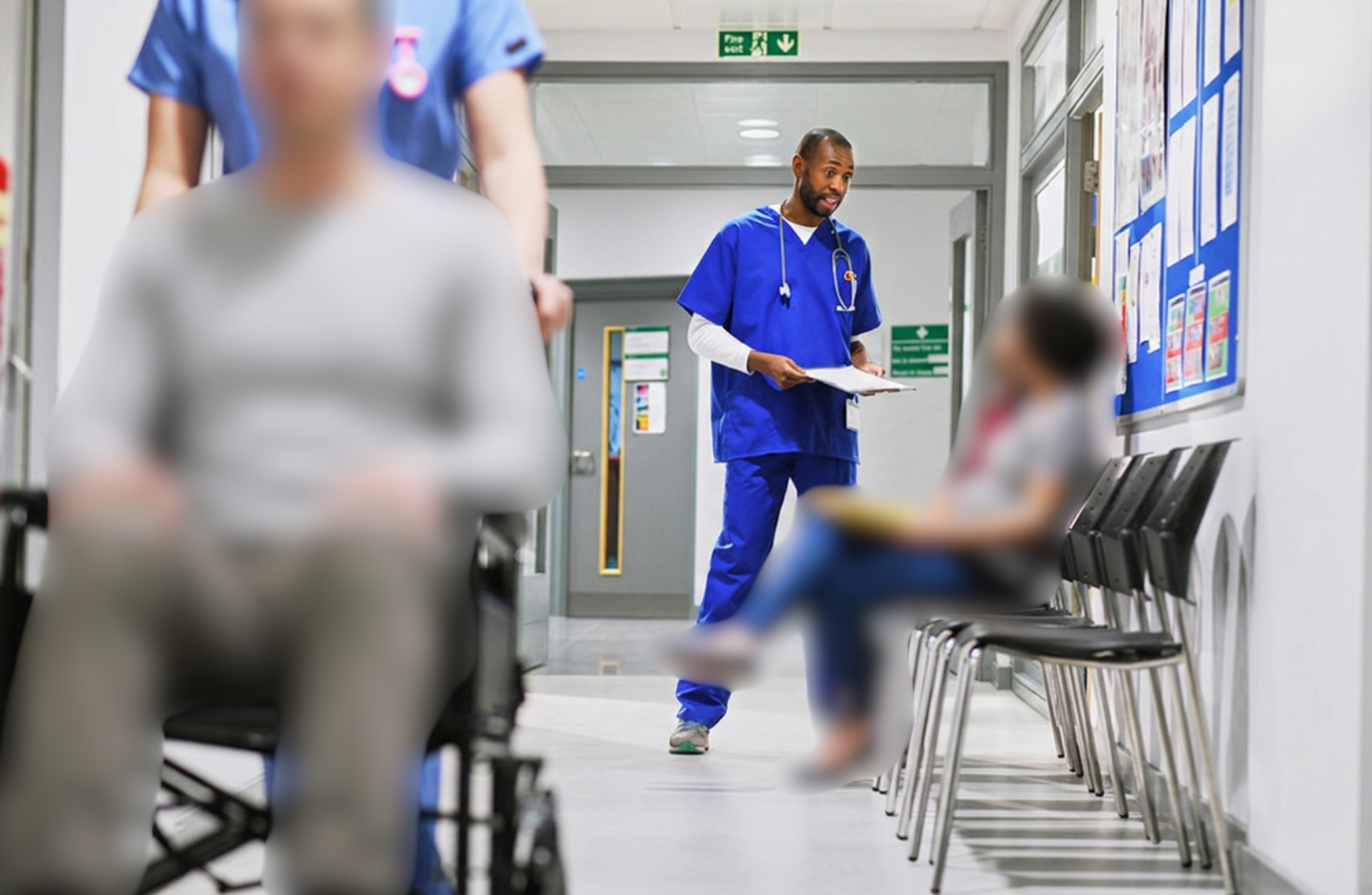 A hallway of a hospital with one nurse pushing a patient in a wheelchair, and a doctor in blue scrubs talking to two people sitting down in chairs. All the patients are blurred using privacy ai.