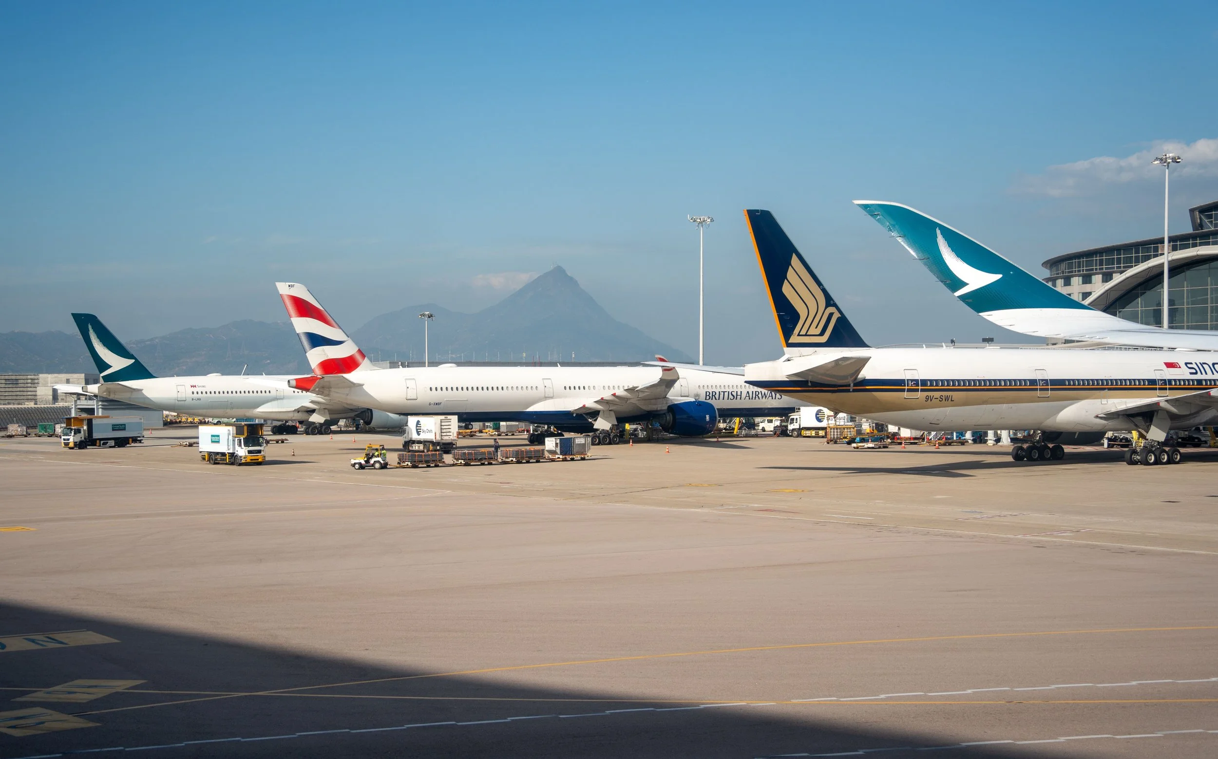 Several commercial airplanes parked at an airport tarmac with ground support vehicles nearby. The background shows mountains and a clear blue sky.